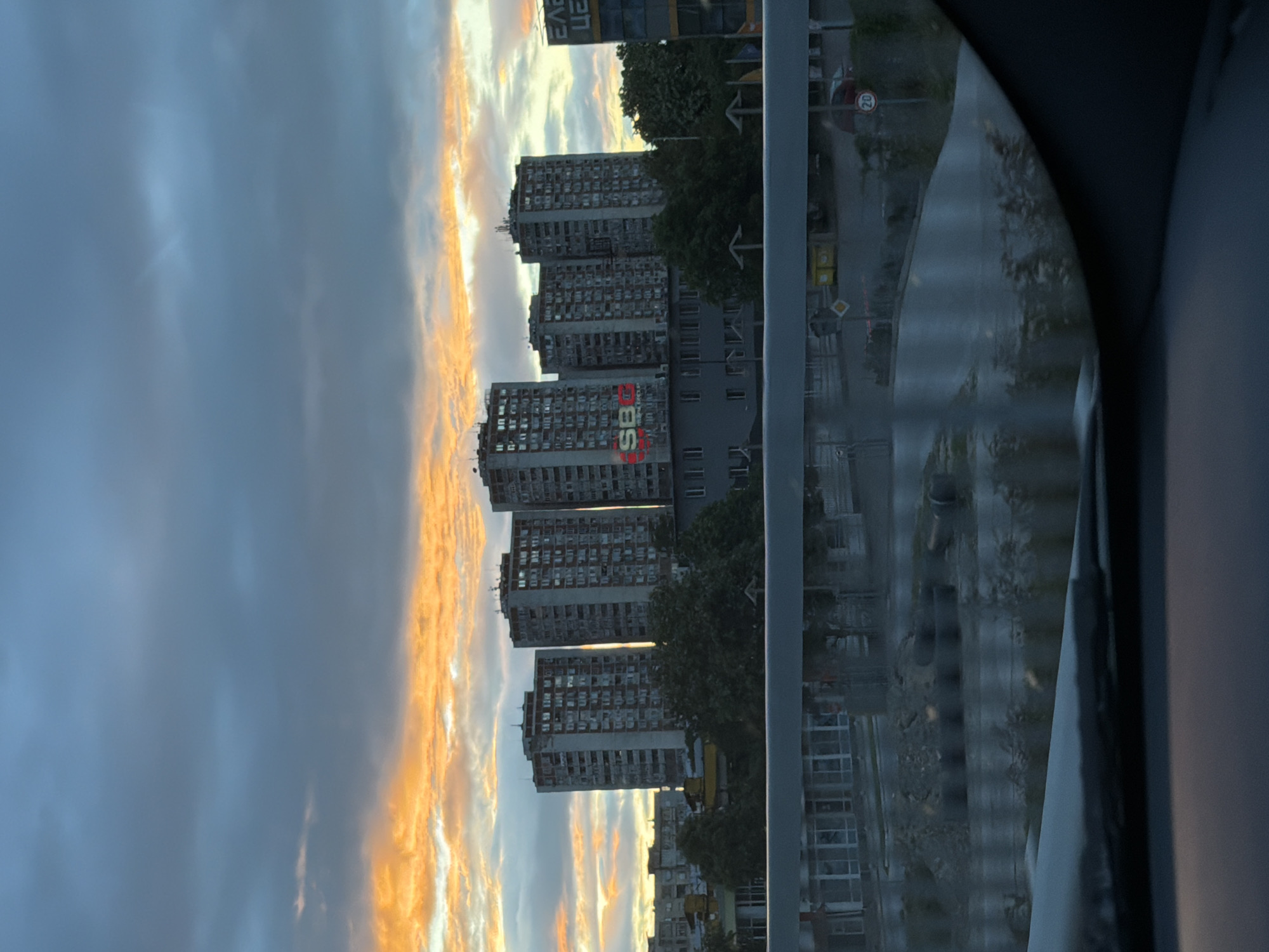 The image shows a view of the Trakiya neighborhood in Plovdiv, Bulgaria, taken from inside a vehicle, likely a car. The buildings in the foreground are several high-rise apartment blocks, characteristic of socialist-era urban planning. The "SBC" logo is visible on one of the buildings. The sky is filled with clouds, with a band of orange and yellow hues near the horizon, indicating sunrise or sunset. The road and surrounding environment appear to be wet, suggesting recent rain. Other buildings and infrastructure are present along the road. A "20" speed limit sign is also visible. The image is framed by the vehicle's window, with part of the car's interior visible on the right side of the frame.
