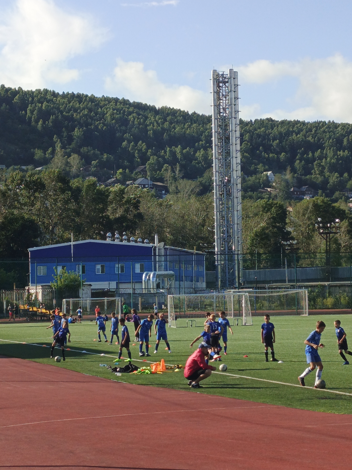 The image depicts a group of young boys engaged in a soccer practice on a bright, sunny day at a sports complex in Zlatoust, Russia. The field, covered in green turf, is marked with white lines and dotted with orange cones, indicating training drills. Several boys, dressed in blue soccer uniforms, are actively participating, some kicking soccer balls, while others appear to be running or observing. One boy in a red jacket and black shorts is crouching on the field, possibly coaching or supervising.
In the background, a long, blue building with white-trimmed windows and a metal roof structure is visible, likely part of the sports facility. Towering behind the building is a tall, silver, lattice-like structure, possibly an industrial chimney or communication tower, characteristic of Zlatoust's industrial landscape. A densely forested hillside rises behind the structure, with scattered houses visible among the trees, suggesting a semi-urban or suburban setting. The sky is a clear blue with scattered white clouds. The overall scene is one of a community sports event or training session set against the backdrop of Zlatoust's natural and industrial environment.