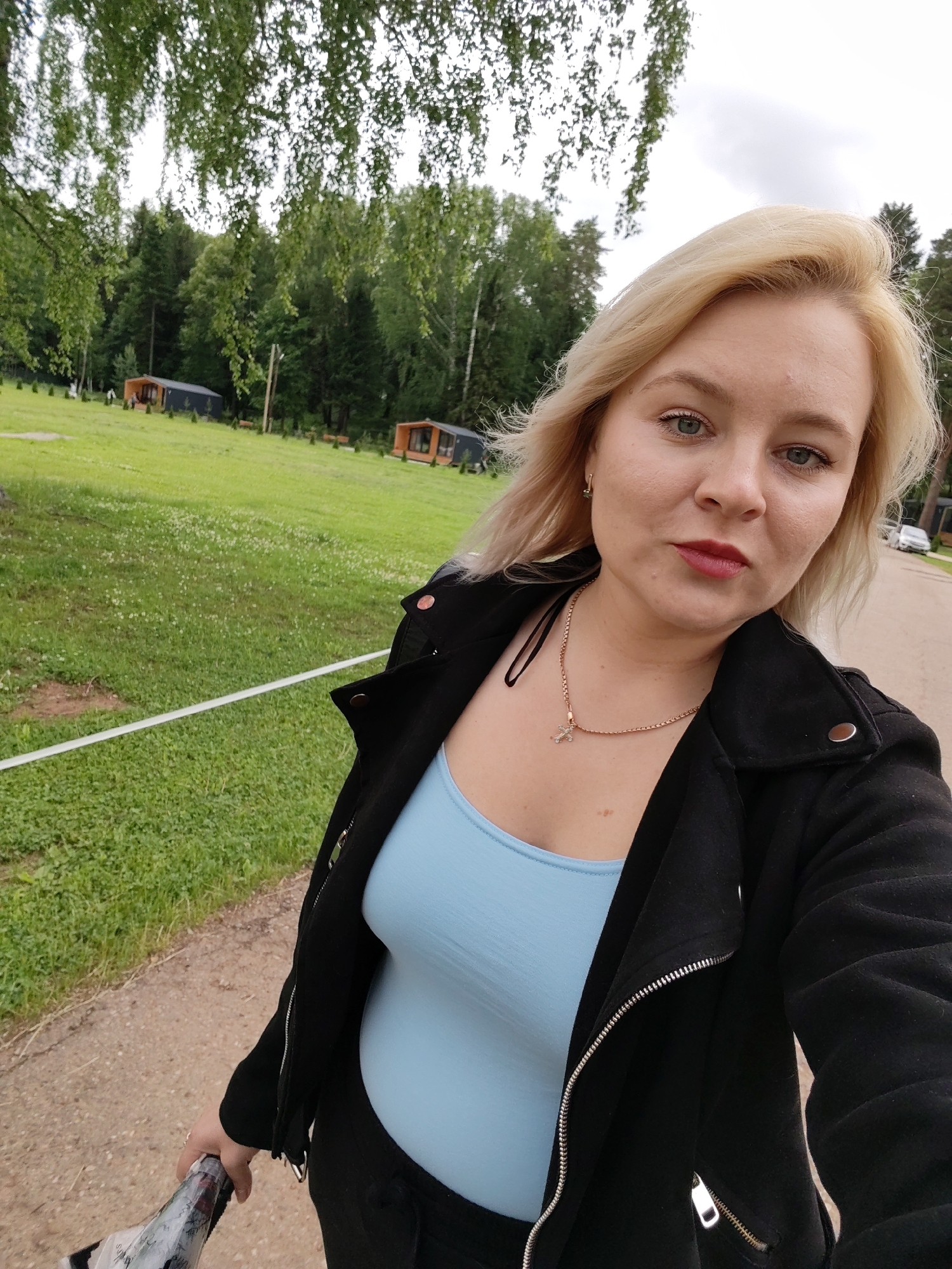 A blonde adult female is depicted in a close-up selfie outdoors, against a natural backdrop in the city of Doma 1096 Km, Russia. She has light-colored eyes, fair skin, and red lipstick. Her shoulder-length hair is styled with volume around her face. She wears a light blue tank top beneath a black, zippered biker-style jacket and a gold-colored chain necklace with a small cross pendant. A gold earring is visible in her left ear. Her right hand, partially visible at the bottom right, holds what appears to be a rolled-up black umbrella. The woman looks directly at the camera with a neutral expression.
The foreground features a dirt path in the lower left, leading to a green grassy field bordered by a white horizontal barrier. The field contains scattered white flowers and several small, conical evergreen shrubs. In the mid-ground, two distinct modern cabins, dark in color with wooden facades and large windows, are visible in the grass, suggesting accommodation units. A dense forest with both deciduous and coniferous trees forms the background. Overcast skies are visible above the tree line. The upper portion of the image is framed by blurred green leaves hanging from overhead trees. A paved road and a light-colored vehicle are discernible in the far background on the right side.