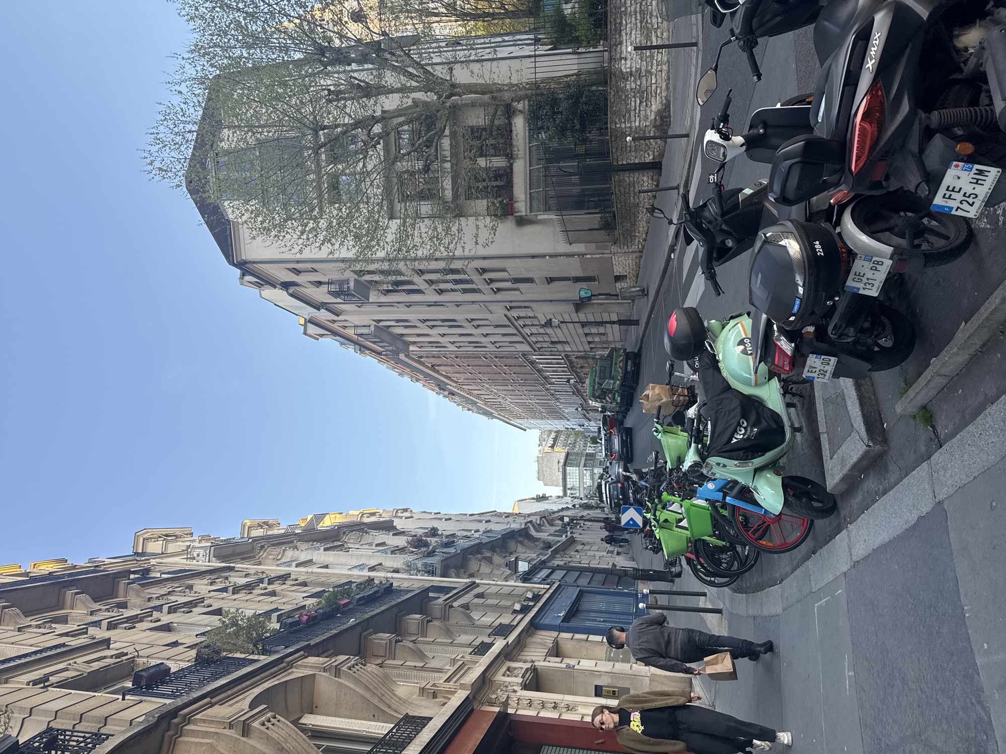 The image shows a street in Paris, France, lined with tall buildings on both sides, typical of Parisian architecture. The sky is clear and blue. Parked along the right side of the street are numerous scooters and motorcycles. Several people are walking on the sidewalk in the lower foreground. The parked motorcycles have license plates showing they are registered in France (FE 725-HM) and Geneva (GE 131-PB, EV 132-OD), hinting at the city's international appeal. Brands "YEGO" and "XMAX" are visible on some of the vehicles. The scene suggests a regular day in the city with people going about their activities, and the scooters parked indicate a popular mode of transportation.