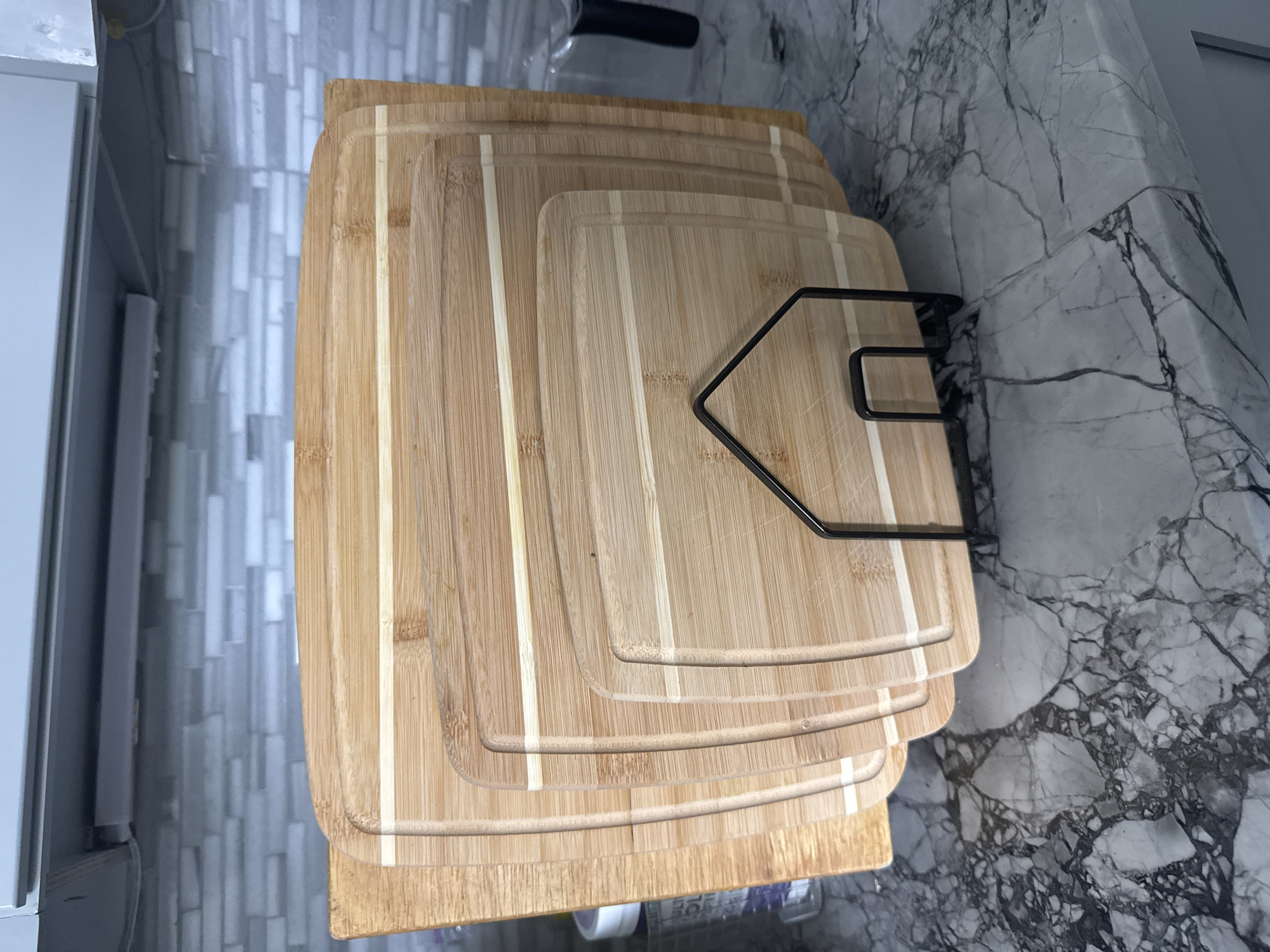 A set of five wooden cutting boards of decreasing size are displayed in a black metal rack on a marble countertop. The cutting boards are made of bamboo and have a grooved edge to catch juices. The background shows a light-colored tiled backsplash and a gray cabinet. The scene is indoors, likely in a kitchen, and there are no people present. The image is taken from a medium-high angle, looking down slightly on the cutting boards.
