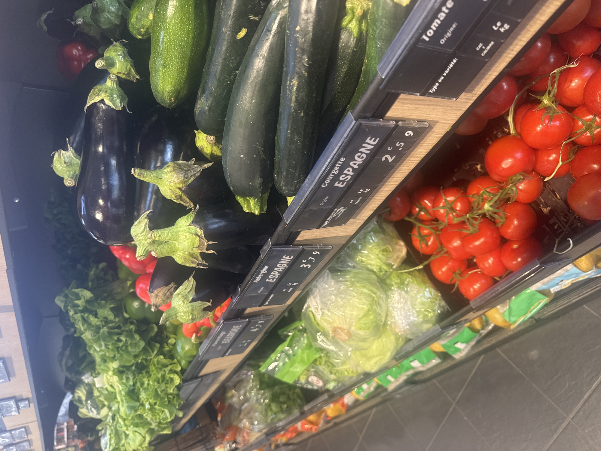 The image shows a produce section in a store, likely a supermarket. The produce is arranged in bins with price tags indicating origin and price per kilogram. Visible produce includes tomatoes on the vine, zucchini, eggplant, and lettuce. Some of the produce has packaging. The price tags indicate that some of the produce originates from Spain and Belgium. The specific city and location are not discernible from the image.