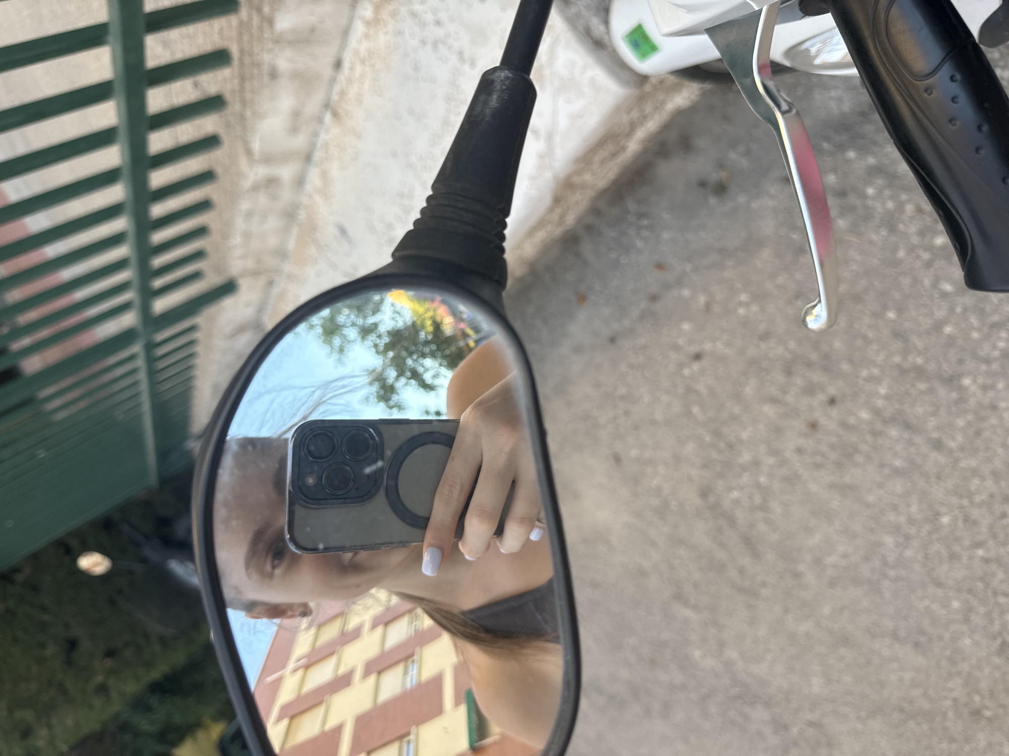 A young woman is reflected in a motorcycle's rearview mirror, holding a smartphone to take a selfie. She has light skin, brown hair pulled back, and is smiling. She is wearing a dark tank top and has light purple nail polish. The background of the reflection shows a building with orange and white stripes, trees, and a blue sky. The motorcycle's handlebars and a brake lever are visible in the foreground. The image is taken from the perspective of someone sitting on the motorcycle. The environment appears to be outdoors in a city.