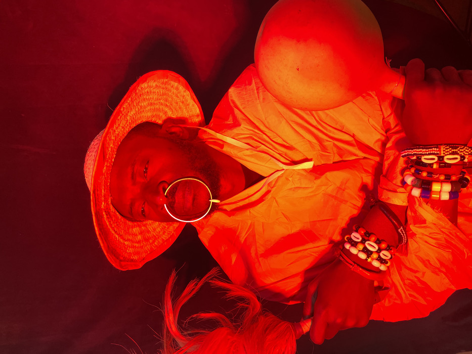 The image depicts an adult male in the city of Jos, Nigeria, illuminated by intense red light. The subject, positioned from the waist up, has dark skin and a short beard, looking upwards and to the right. He wears a wide-brimmed, woven straw hat and an orange-hued collared shirt. A large, circular, golden-toned nose ring extends from his septum to his upper lip. Both wrists are adorned with multiple beaded bracelets; the left features cowrie shells and various beads, while the right displays colorful geometric patterns. In his left hand, he holds a ceremonial whisk or fly-whisk with light-colored fibers. His right hand holds a large, light-colored gourd. The background is uniformly dark, emphasizing the red-lit figure and objects.