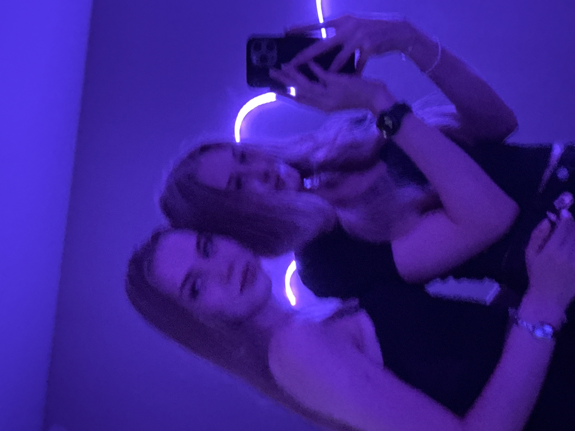Two young women are taking a selfie in a room illuminated by purple lighting. The woman on the left is looking towards the camera, while the woman on the right is holding a smartphone and taking the picture. Both women have long, blonde hair and are wearing dark-colored clothing. A neon light, possibly shaped like a heart, is visible in the background. The room appears to be a bedroom or a personal space, with a plain wall behind them. The overall atmosphere is casual and intimate. There is no information in the image to determine the location in the city of Perm, Russia.