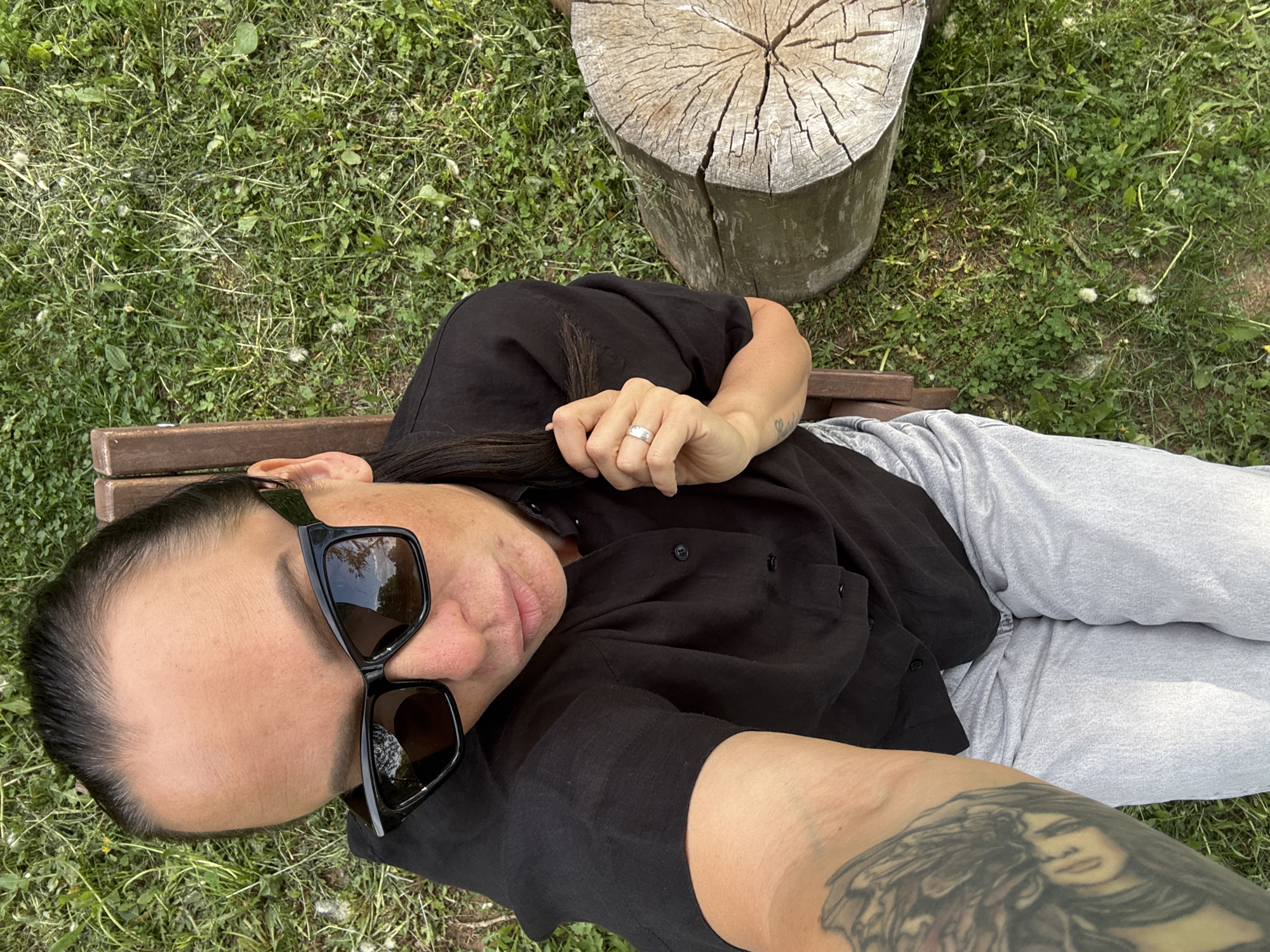 A woman lies on a wooden bench in a grassy area, possibly a park, with a tree stump visible nearby. She wears black sunglasses, a black button-down shirt, and light grey pants. A ring is visible on her left ring finger, and a tattoo is present on her arm closest to the camera. The location is in Chaikovsky, Russia.