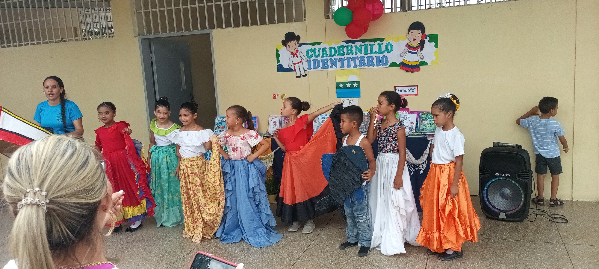 A group of children and an adult are posing for a picture in front of a wall decorated with balloons, posters, and signs written in Spanish. The children, dressed in colorful costumes with a traditional Latin American flair, are smiling and looking towards the camera. The adult woman on the left side of the image is wearing a blue t-shirt and holding a partially obscured triangular object covered with the colors of the Venezuelan flag. To the right of the group, a black Aiwa speaker stands next to a boy in a striped shirt, who is leaning against the wall. The background features a beige wall with a metal gate above a doorframe on the left and several posters and signs reading "Cuadernillo Identitario" and "1 Grado C", suggesting a school event or performance. A woman with blond hair in a ponytail can be seen in the foreground, appearing to be taking a photo with a smartphone.