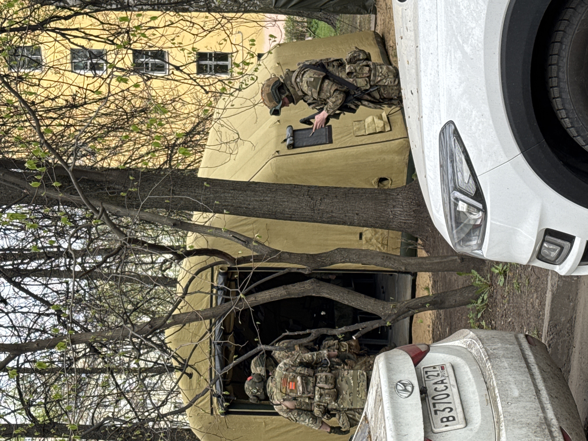 The image shows two soldiers in camouflage uniforms positioned near a light-colored tent structure situated between two parked white cars, one possibly a hatchback and the other, partially visible, an SUV. One soldier is near the front of the tent, seemingly interacting with a screen or device attached to the tent's exterior. The license plate of one car is visible, reading "B 370 CA 27 RUS," indicating a Russian origin. The environment is urban, with trees in the foreground obscuring some of the view. A yellow building with windows is visible in the background. Saint Petersburg is not explicitly mentioned in the image and cannot be confirmed.