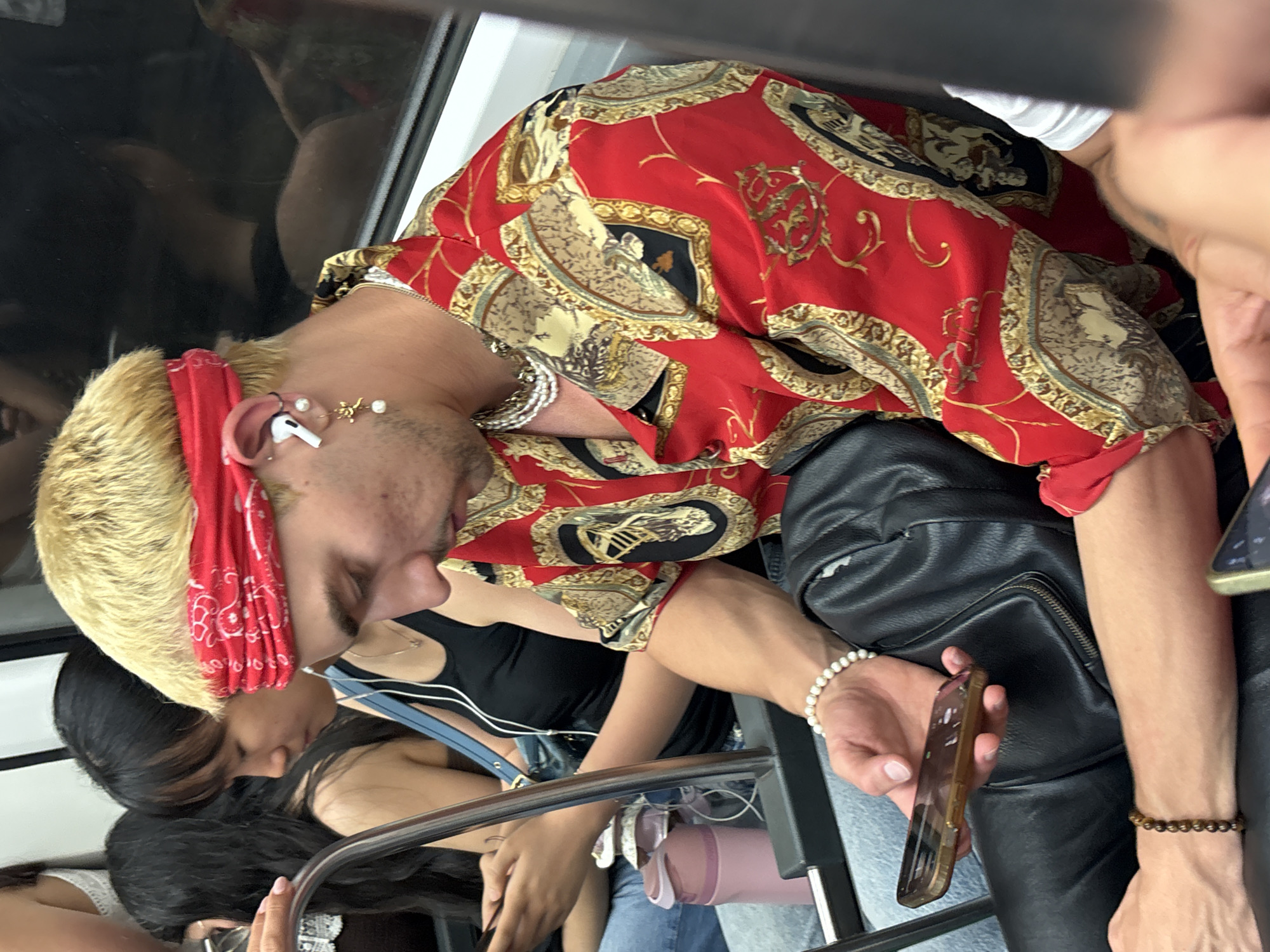 In l'Hospitalet de Llobregat, Spain, a blonde-haired individual wearing a red bandana and a patterned red shirt is seated on public transportation, possibly a train, holding a smartphone. To their left, a woman with dark hair is also seated, looking down at her phone. Another woman with dark hair is seated behind them. The environment appears to be an indoor setting with a window and seating.