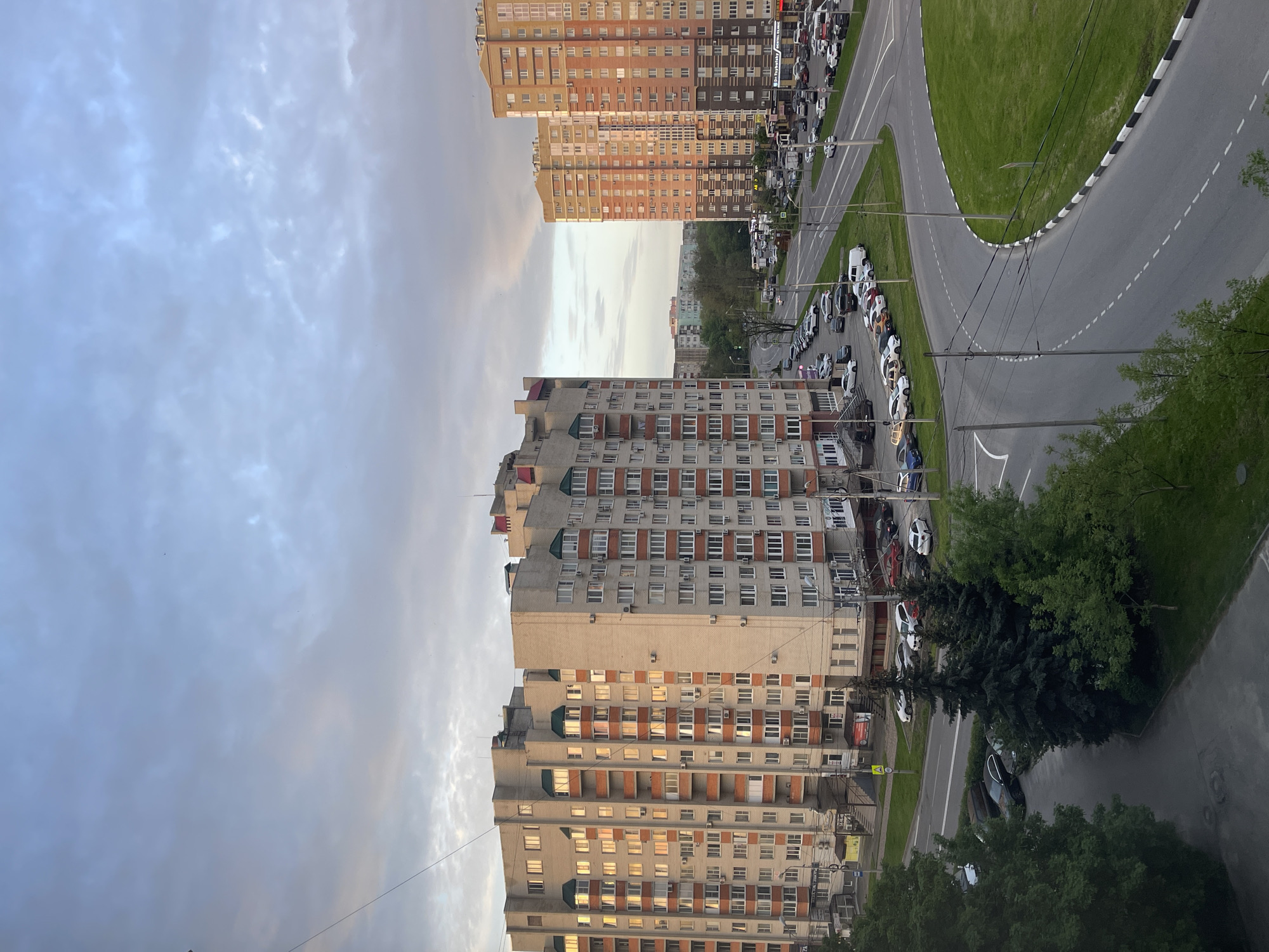 The image shows a view in Stavropol, Russia, featuring residential buildings, a street with parked cars, and a roundabout. The foreground has a tall, beige apartment building with rows of windows and orange accents. A line of parked vehicles is visible on the street beside the building, along with trees and road markings. Beyond the roundabout, a second, taller residential building in shades of orange and brown rises up, similarly lined with rows of windows. The sky occupies a significant portion of the image, displaying a mix of clouds and some clear blue areas, suggesting either dawn or dusk.
