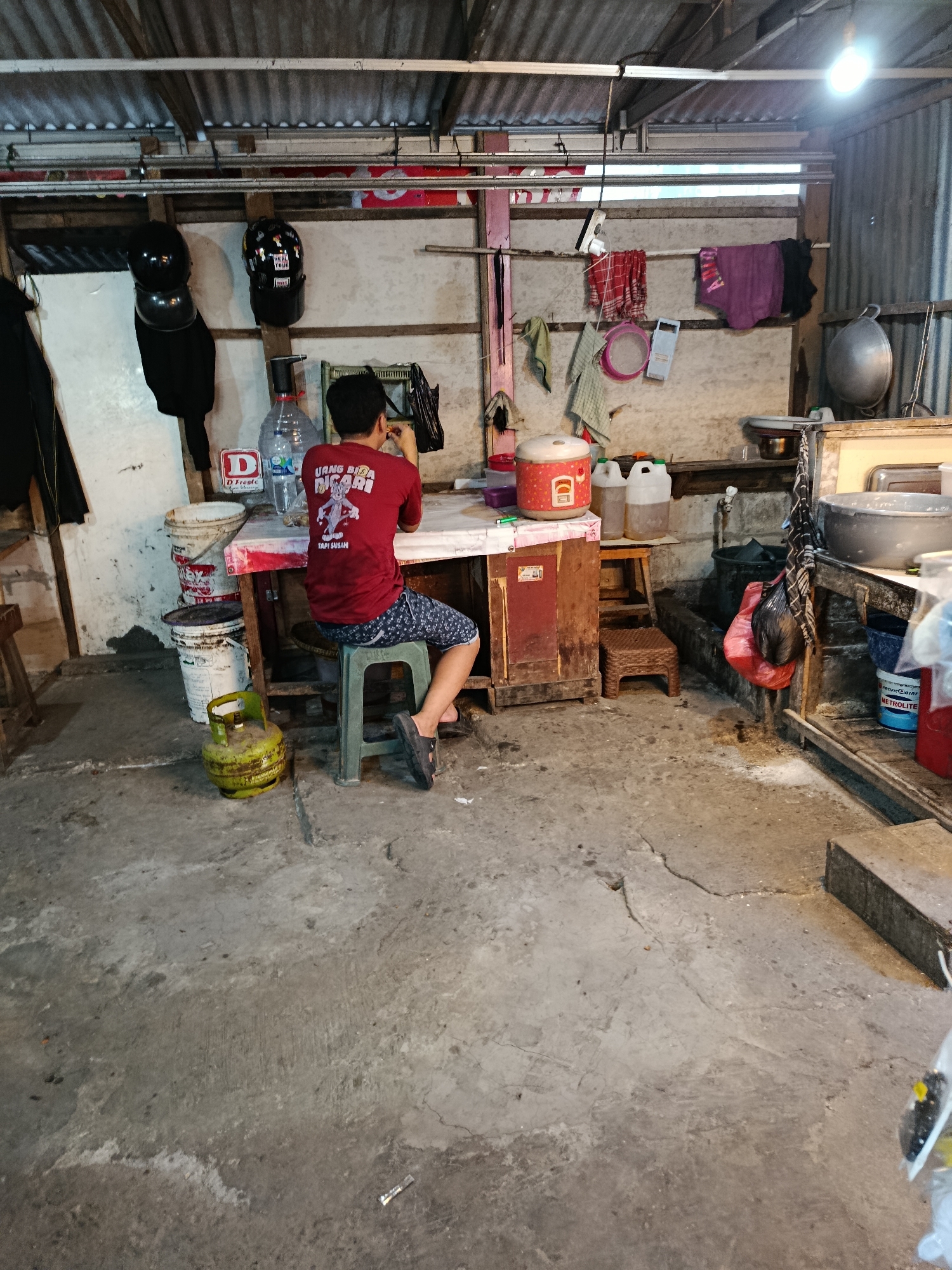 A young male, seated on a stool with his back to the camera, is positioned at a makeshift table in what appears to be an outdoor or semi-enclosed kitchen area. He wears a red t-shirt with a cartoon character and text on the back, and patterned shorts. His hands are near his mouth, suggesting he might be eating or engaged in some activity with his hands.
The environment is cluttered and utilitarian, typical of a simple, functional workspace. The ceiling is corrugated metal, supported by exposed wooden beams. The walls are rough and uneven. Various items are visible, including several large plastic buckets, one of which contains a gas cylinder. A water dispenser with a large bottle is also present. The table itself is a red-topped surface with a wooden cabinet underneath. On the table, a rice cooker and several plastic containers are visible. Hanging on the wall are various items of clothing, towels, and some miscellaneous objects. The floor is concrete, showing signs of wear and tear. In the background, large metal basins and other kitchenware are arranged.
The scene likely depicts a humble food stall or a home kitchen in a less developed area. The presence of the gas cylinder and the overall rustic aesthetic suggest a practical, resource-conscious setting. The lighting appears to be artificial, likely from a bulb hanging overhead. The image captures a candid moment of everyday life in a working-class environment. Given the context of Balaraja, Indonesia, this scene is representative of the many small businesses and residential areas found in such locations, characterized by resourcefulness and a focus on practical needs.