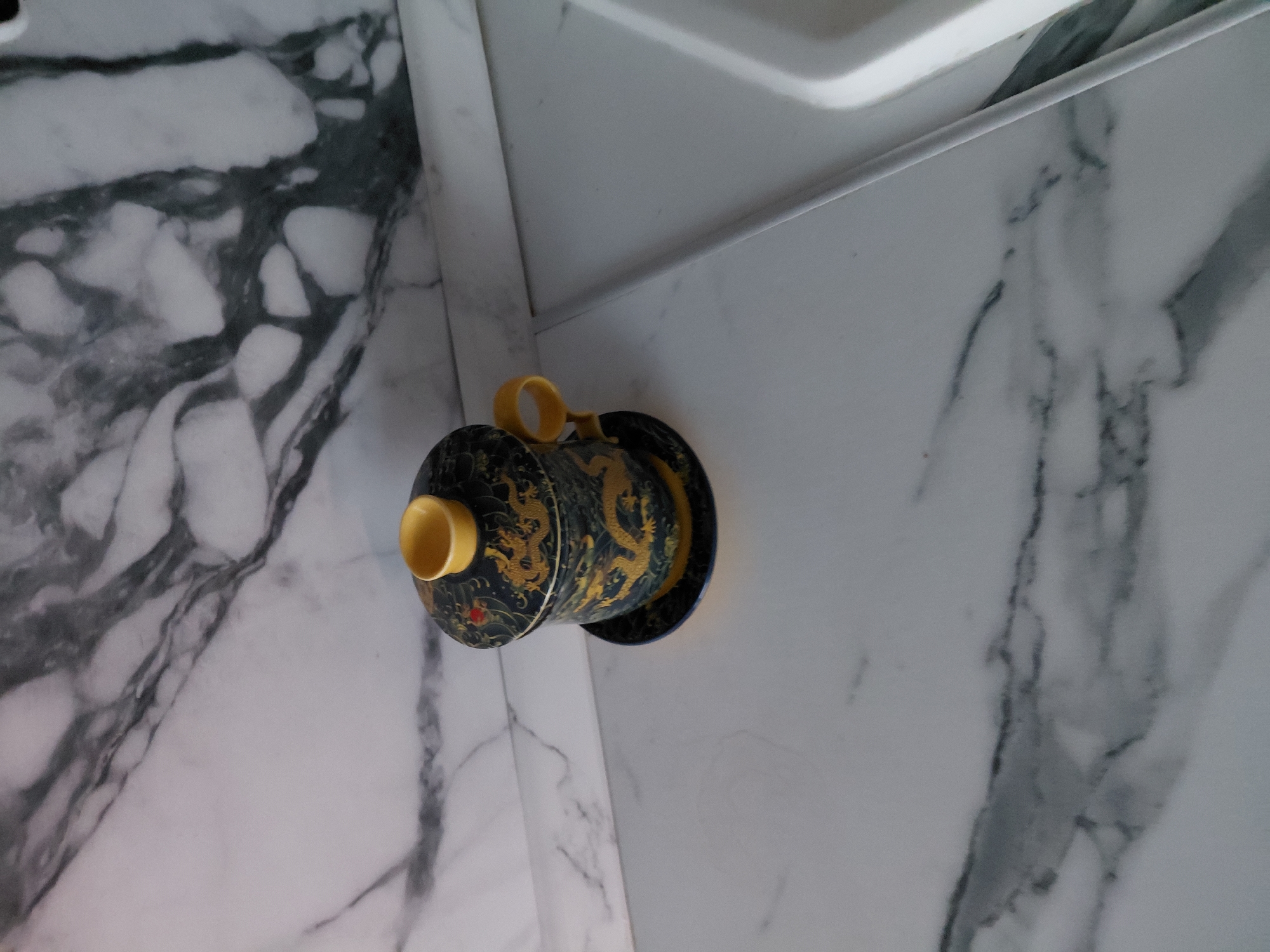 The image displays a decorative cylindrical object, possibly a tea caddy or urn, featuring intricate golden dragon designs on a dark, likely black or deep blue, background. The object has a yellow or golden handle-like detail on its lid and a matching rim at the base. It is placed horizontally on a glossy white surface that resembles marble, characterized by prominent grey and black veining. In the background and upper right, white, possibly ceramic or plastic, edges or fixtures are visible, suggesting an indoor setting such as a kitchen, bathroom, or utility room. The lighting is bright and even, indicative of an indoor environment during daytime, though the specific time of day or outdoor weather cannot be determined. No activity or event is taking place, and no people or visible text are present. The setting is an interior space in Nadym, Russia.