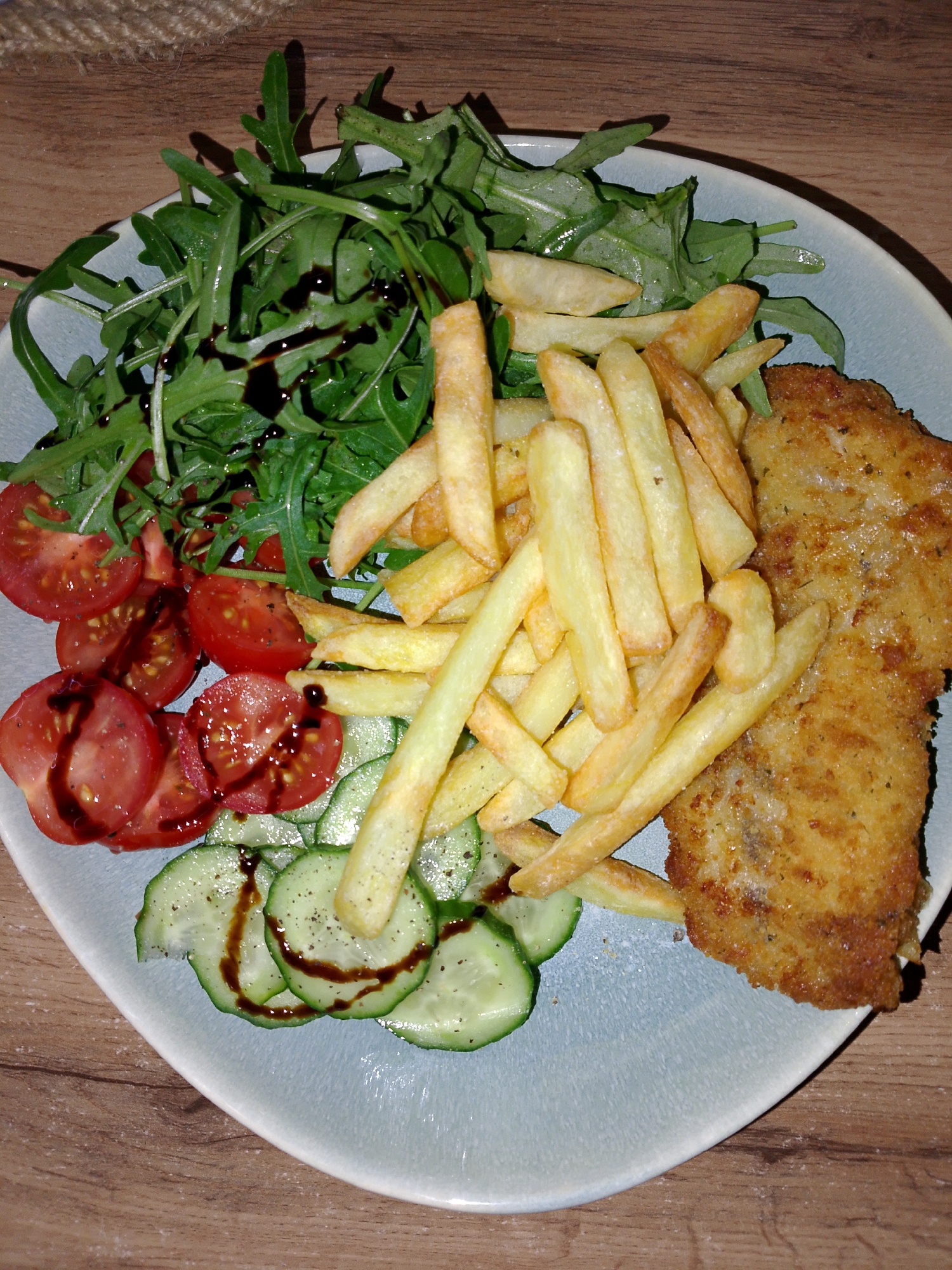 The scene depicts a prepared meal presented on a light-colored, possibly ceramic, plate, positioned on a wooden surface within Izhevsk, Russia. The meal comprises a large, golden-brown breaded cutlet or schnitzel occupying the right side of the plate. Adjacent to it, a pile of golden french fries is centrally located. To the left, a fresh salad portion includes a cluster of green arugula leaves at the top, followed by several slices of red tomato below them, and overlapping slices of green cucumber at the bottom. A dark, viscous dressing is drizzled across the arugula, tomatoes, and cucumbers, with visible black pepper flakes on the cucumber slices. The wooden surface exhibits grain patterns, and a portion of a woven texture, possibly a placemat, is visible in the upper left background. No individuals or specific actions are depicted.