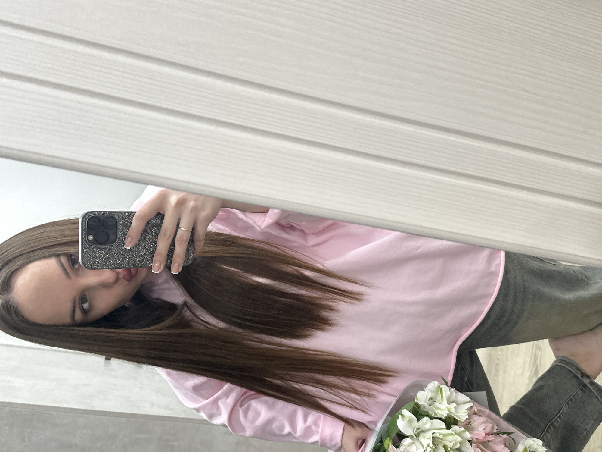 A female subject is depicted taking a mirror selfie indoors. The specific location within Chaikovsky, Russia, cannot be determined from the visual evidence. The woman has long, straight brown hair, styled with a center part. She is wearing a light pink, long-sleeved collared shirt and faded grey jeans. Her right hand, adorned with a gold-colored ring on the ring finger and French-tipped fingernails, holds a smartphone with a dark, glittering case, partially obscuring her face. Her lips are pursed in a 'kiss' gesture. Her gaze is directed towards the phone screen. In her left arm, she holds a bouquet of white and pink alstroemeria flowers, wrapped in transparent material. Her left foot, bare, is partially visible. The immediate background on the left consists of a light grey wall, possibly tiled or textured. On the right, a light-colored door or paneled surface with vertical lines is visible.