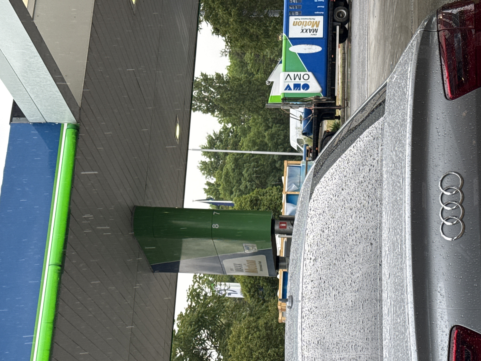 The image depicts a rainy scene at an OMV gas station. A silver Audi car is parked at a gas pump. The gas pump displays prices for various fuels. The gas station is located in Bulgaria.