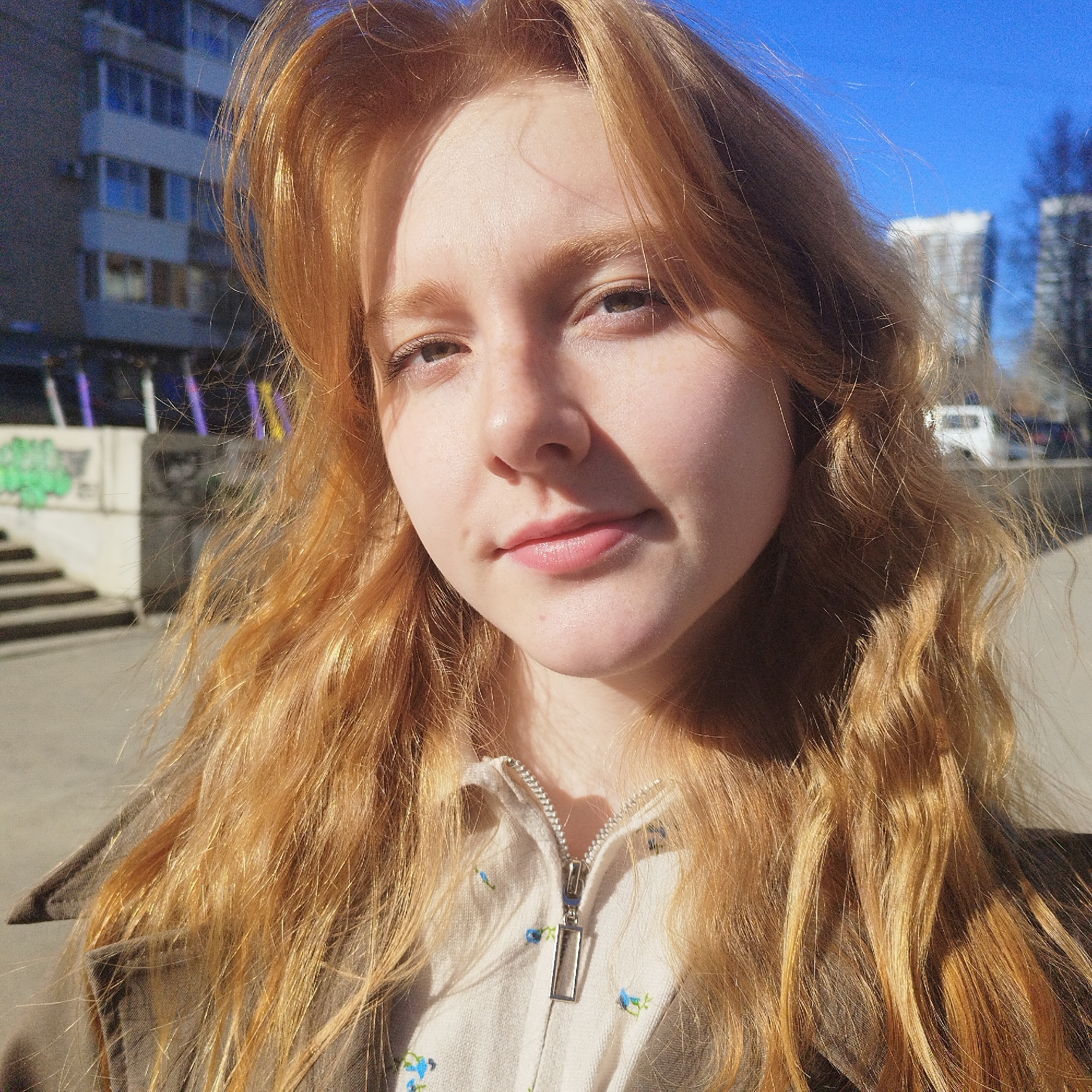 A close-up outdoor portrait features a young woman with long, wavy, light auburn hair, which is brightly illuminated by direct sunlight. She has fair skin with subtle freckles and greenish-brown eyes, and looks directly at the camera with a calm, subtle smile. She is wearing a light-colored, zip-front top adorned with small blue floral embroidery, layered under a darker, possibly brown or olive, jacket or coat.
Strong sunlight casts clear shadows on the left side of her face and neck, accentuating the texture and vibrant color of her hair. The background indicates an urban setting on a clear, sunny day. To the left, concrete steps or a wall are visible, featuring indistinct graffiti. Multi-storied buildings can be seen in the distance under a bright blue sky. A white vehicle is partially visible on the right side, further back. The scene depicts a bright day in an urban environment, with prominent natural lighting. No decipherable text is visible.