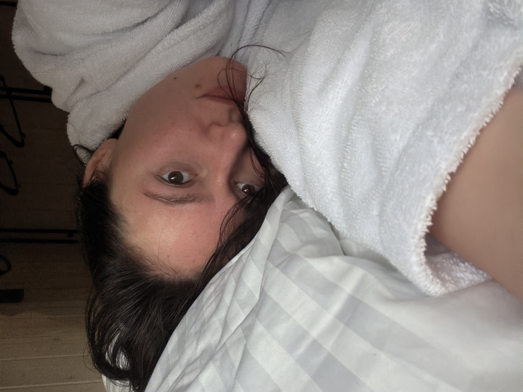 A close-up, overhead shot captures a person lying down, looking directly into the camera. The subject has dark hair, which appears slightly damp or messy, framing their face and resting on a white pillow. Their dark eyes are open and focused. They are wearing a plush, white terry cloth bathrobe, which is visible wrapped around their shoulders and partially covering their body. The bathrobe and the white pillow feature subtle striped patterns, adding texture to the scene.
The lighting in the image is dim, suggesting an indoor setting, possibly a bedroom during morning or evening hours. A dark background element is visible to the upper left, while faint hints of wooden floorboards appear along the far left edge, further indicating an interior space. The person's expression is neutral and calm. There is no visible text in the image. The overall atmosphere is intimate and quiet.