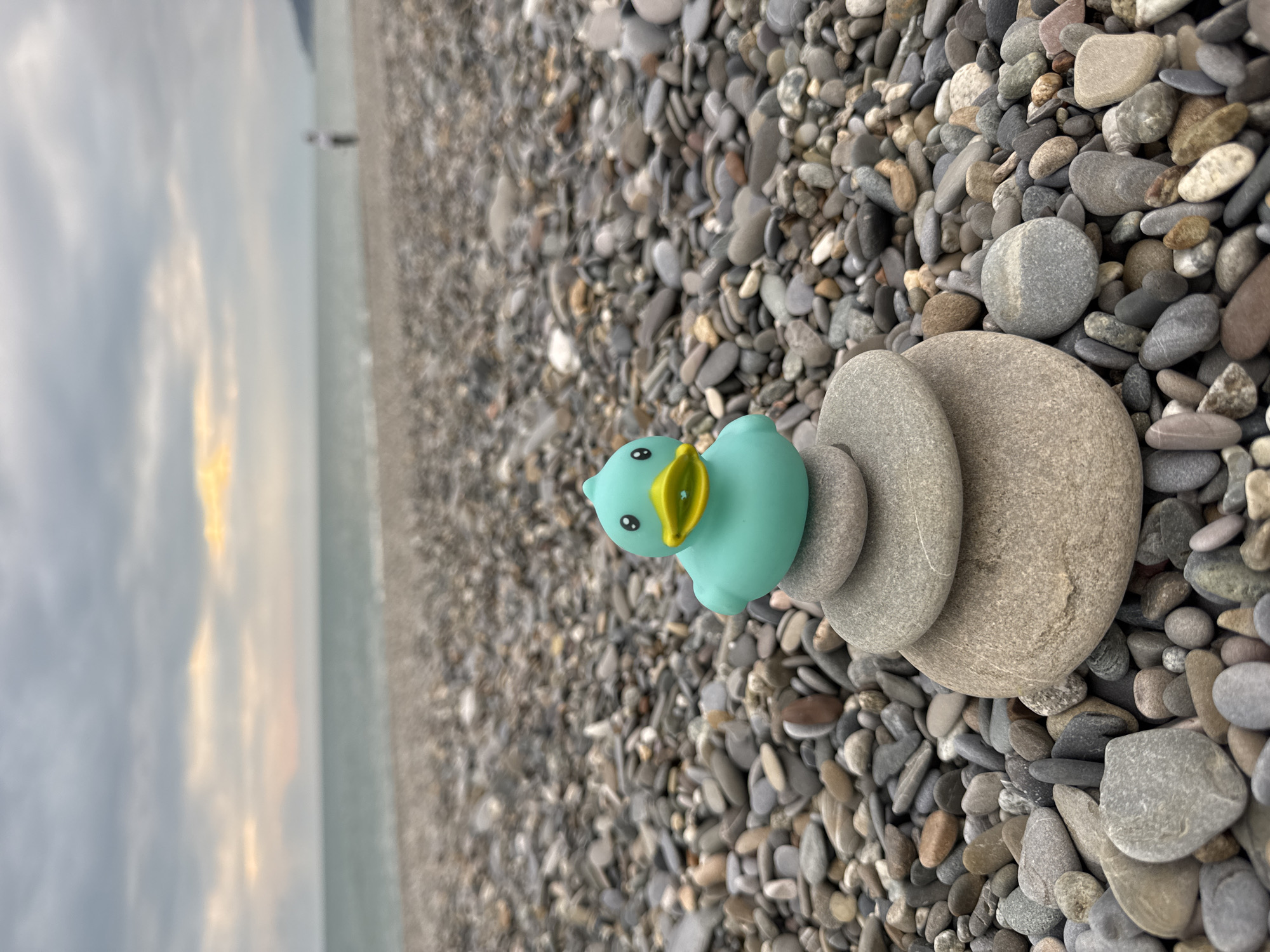 The image captures a blue rubber duck resting atop a stack of three gray stones on a pebble beach in Sochi, Russia. The stones are stacked from largest to smallest. The beach is composed of small, smooth, gray and tan pebbles that stretch into the distance. In the background, the sea meets a sky filled with gray clouds reflecting the light of the setting sun. A figure is visible in the distance near the waterline.