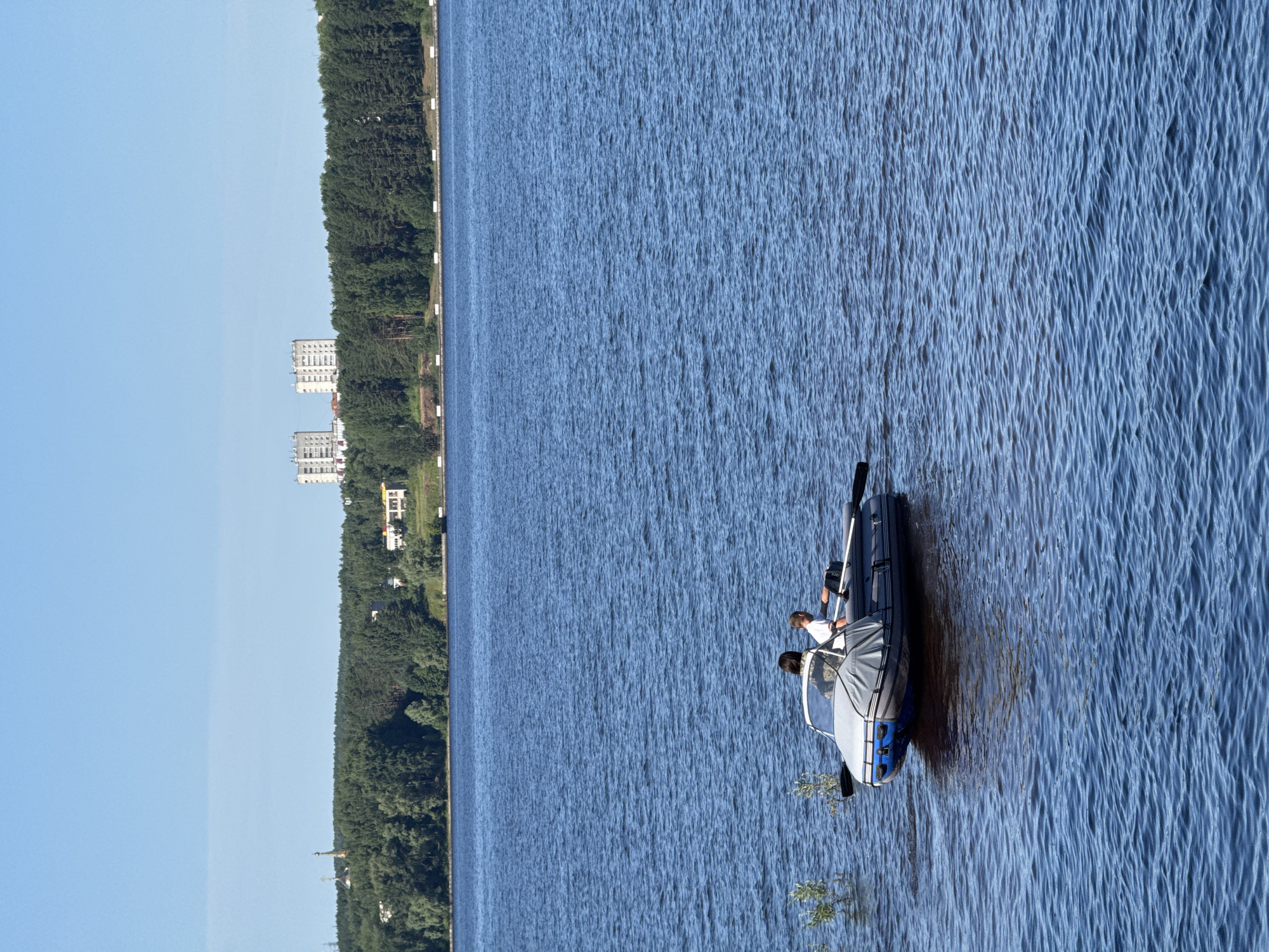 In Chaikovsky, Russia, on a clear, sunny day, a wide, blue body of water is visible. The water is slightly choppy with small waves. In the middle ground of the image, a small, inflatable boat is navigating the water. Two individuals are present in the boat; one person is seated and appears to be steering or rowing, while another person is seated behind them. The boat has a canopy covering part of it. In the background, a dense line of green trees forms the shoreline, with several multi-story buildings visible beyond the trees. The sky above is a clear, light blue.