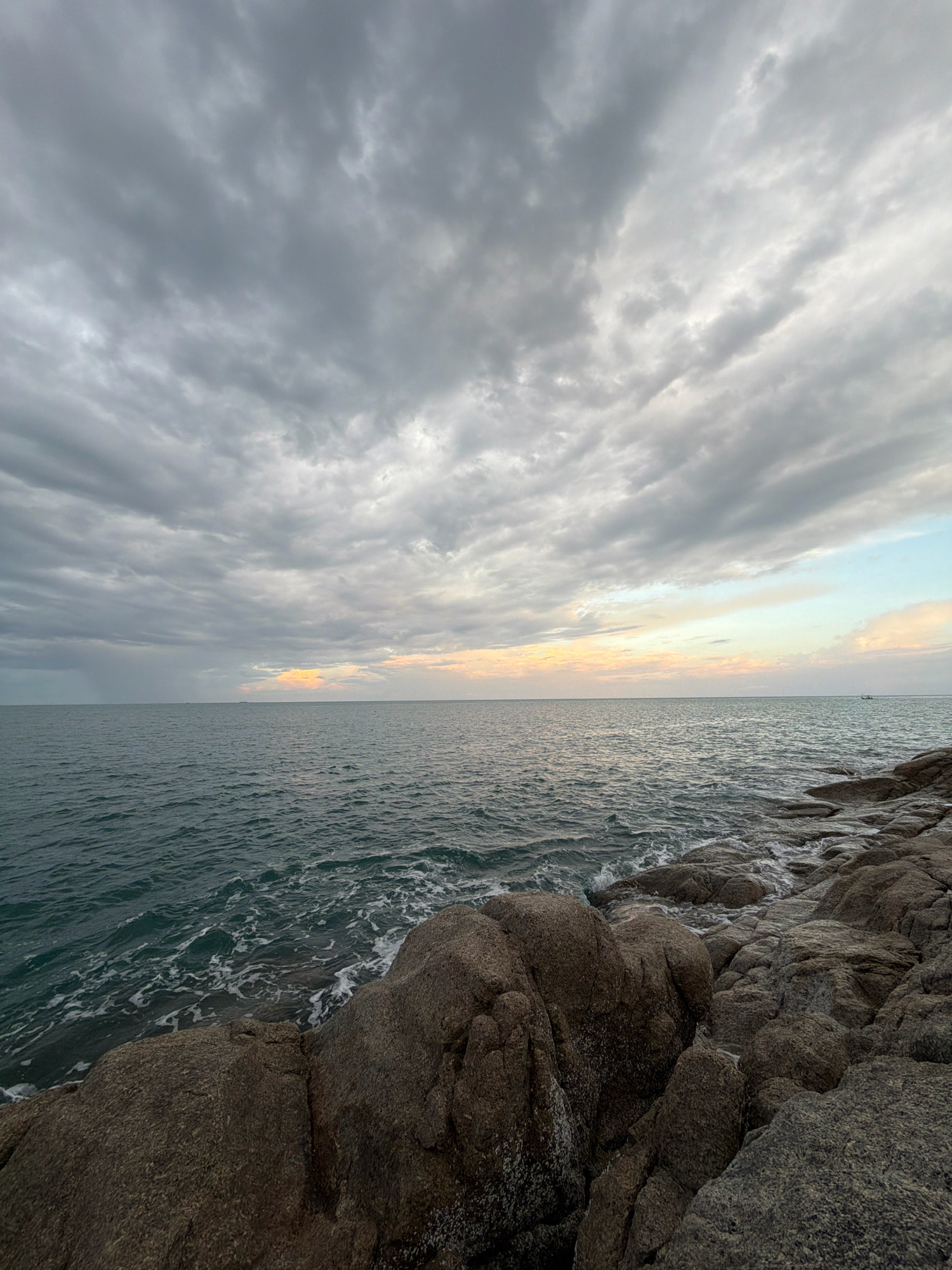 This is a wide-angle, low-angle shot of a rocky coastline and the sea under a cloudy sky. The foreground is dominated by large, grey, weathered rocks, some of which are partially submerged by the incoming tide. The sea is a deep teal color with white foam washing up on the shore. The horizon line is visible, with a muted, colorful sky above, hinting at either sunrise or sunset. There are faint clouds stretching across the sky, with some appearing darker and more dense than others. In the distance, on the right side of the frame, a small boat is visible on the water. The location is identified as Ko Samui District, Thailand.