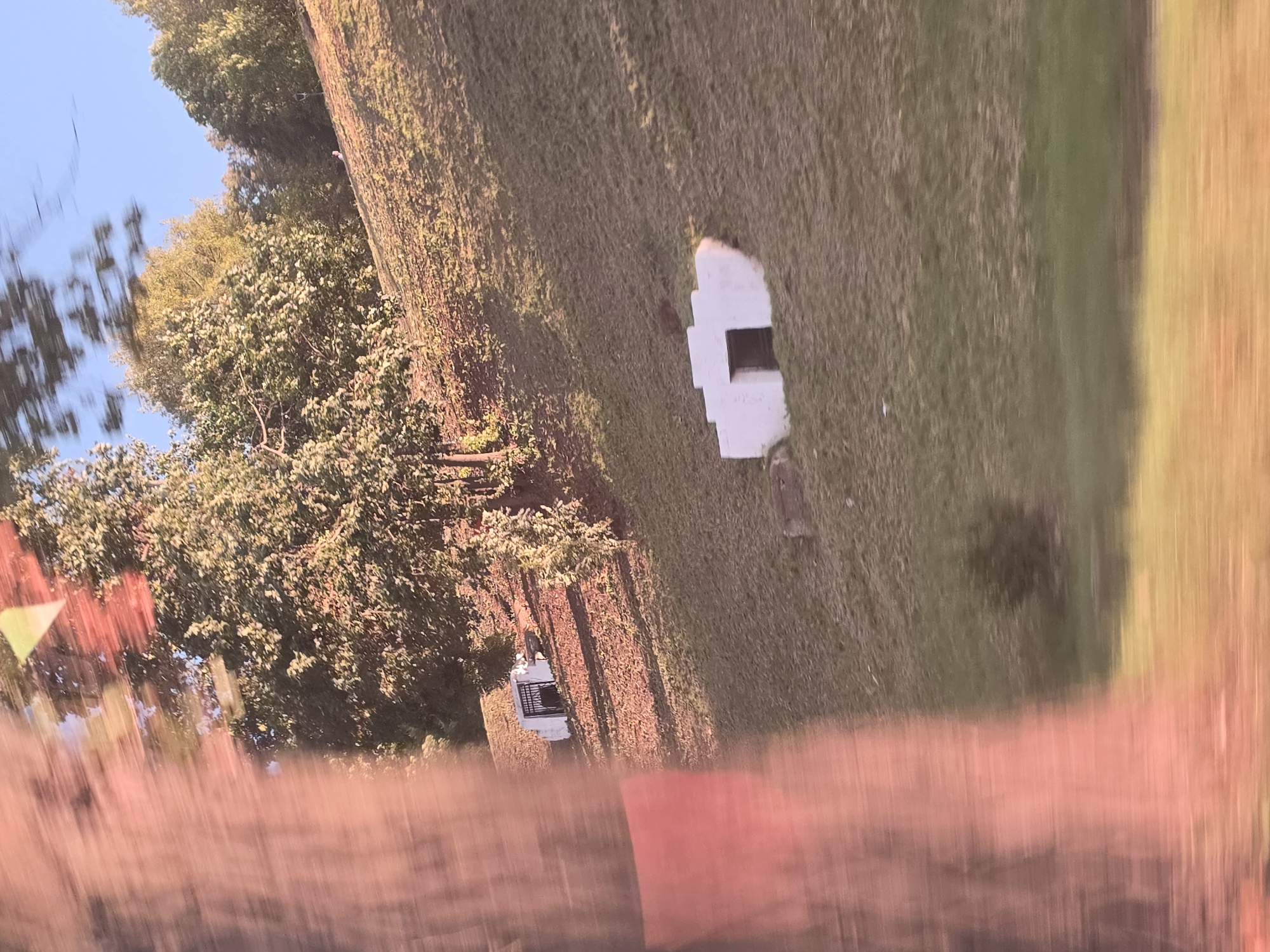The image depicts a hillside with two small, white structures built into it, each with an open doorway. The structure on the left has a black metal gate in its opening. The hillside is covered in grass and fallen leaves, and trees with green foliage are visible at the top of the slope. The sky above is clear and blue. The image appears to be taken from a moving vehicle, as indicated by motion blur in the foreground, particularly on the left side where a large, reddish-brown blurred object, possibly a tree trunk or a sign, is visible. The scene is bathed in warm sunlight, casting long shadows. No people are clearly identifiable in the image. The context of Nizhny Novgorod, Russia, is not visually evident from the image itself.