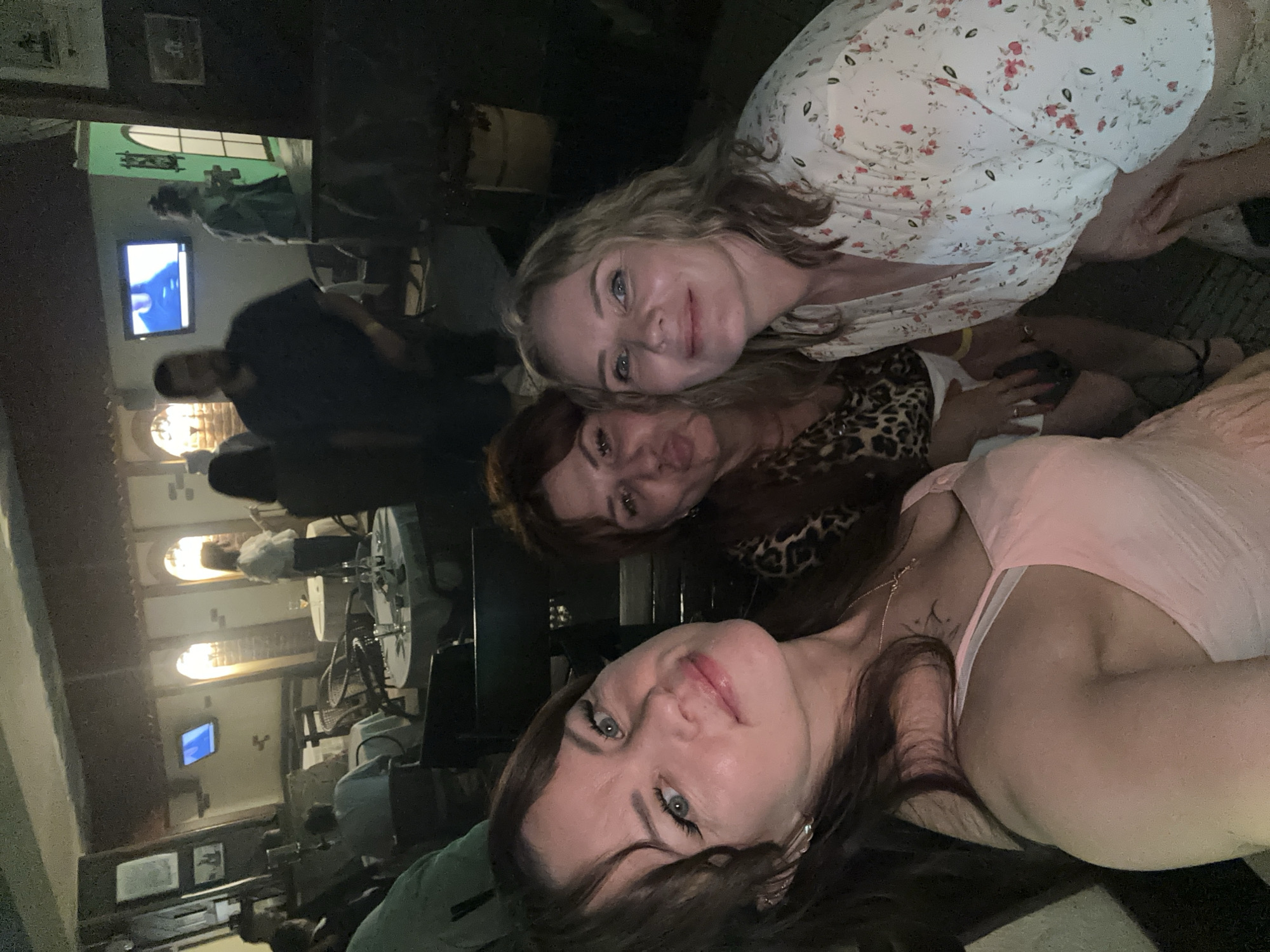 The image depicts an indoor scene at what appears to be a restaurant or bar in Chaikovsky, Russia. Three women are prominently featured in the foreground, posing for a selfie. The woman on the left, with dark hair and blue eyes, is wearing a light pink dress and looking directly at the camera. To her right is a woman with brown hair, making a kissing face and wearing a leopard-print top. The third woman, on the far right, has blonde, wavy hair and is smiling, dressed in a floral print garment.
In the background, other patrons and restaurant elements are visible. Tables with white tablecloths and chairs are arranged throughout the space. A television screen is mounted on the wall, displaying an image. The lighting is dim, contributing to an evening or nighttime ambiance. The architectural style of the establishment, with exposed wooden beams and arched doorways, suggests a rustic or traditional theme. The overall setting appears to be a social gathering or outing.