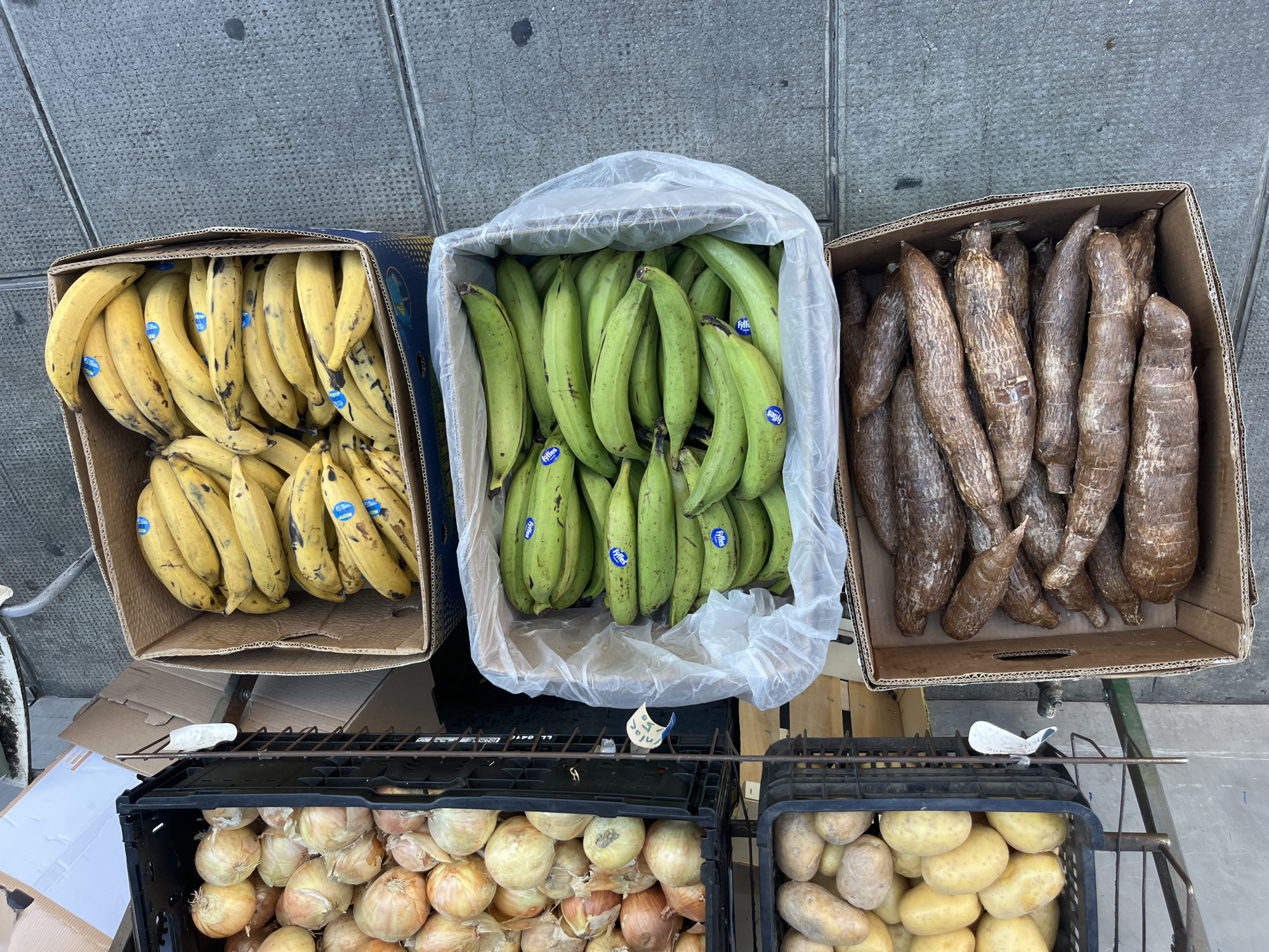 The image captures a street-level view of a produce stand, likely in Geneva, Switzerland, given the contextual information. In the foreground, stacked crates and boxes display a variety of fruits and vegetables. From top to bottom, a large cardboard box is filled with ripe yellow bananas. Below this, a white plastic-lined box contains green plantains. To the left of the plantains, a black plastic crate holds a substantial quantity of yellow onions. Further down, another black plastic crate is filled with potatoes. Finally, at the bottom right, a cardboard box is filled with elongated, brown-skinned tubers, likely yucca or cassava. The ground surface appears to be paved, consistent with a sidewalk or street. No people are visible in the frame. The lighting suggests it is daytime.
