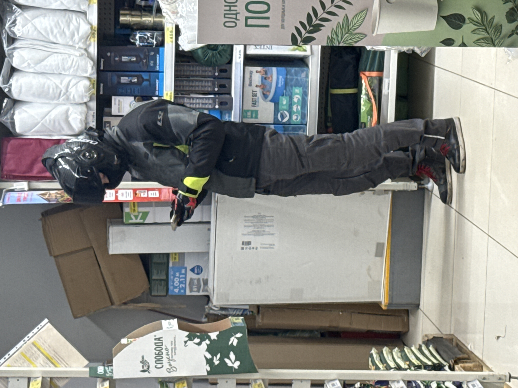 The rotated image depicts a person inside what appears to be a retail store or warehouse. The individual is wearing a black full-face helmet, a dark jacket with bright yellow accents on the sleeves and waist, dark grey pants, and black shoes with prominent red laces. They are standing upright, leaning slightly forward, with their hands held low, possibly looking at items on a lower shelf or inspecting something on the floor.
The setting is an aisle stocked with various products. On the left side, shelves hold stacks of white pillows or quilts packaged in clear plastic. Above these are blue boxes containing what appear to be electronics or small appliances. Further along the aisle, visible behind and to the right of the person, are items related to outdoor recreation, including a box for a "Bestway FAST SET" swimming pool and packaging with the "INTEX" brand name. A large, plain white appliance box is prominent in the mid-ground, surrounded by several brown cardboard boxes. At the bottom left, a box is visible with the Cyrillic text "СЛОБОДА Вкусно" (SLOBODA Delicious). Other visible text includes "ОДНО ПО" (ODNO PO) and "для холодных и го" (for cold and hot...) on a sign above, and dimensions "4.00 M x 2.11 M" on one of the boxes.
The floor is made of light-colored tiles, characteristic of a commercial establishment. The person's attire, particularly the helmet, suggests they may have arrived on a motorcycle or scooter. The scene is indoors, presumably during store operating hours. The context of Yekaterinburg, Russia, aligns with the Cyrillic text visible on the packaging and signage. The activity appears to be shopping or browsing merchandise.