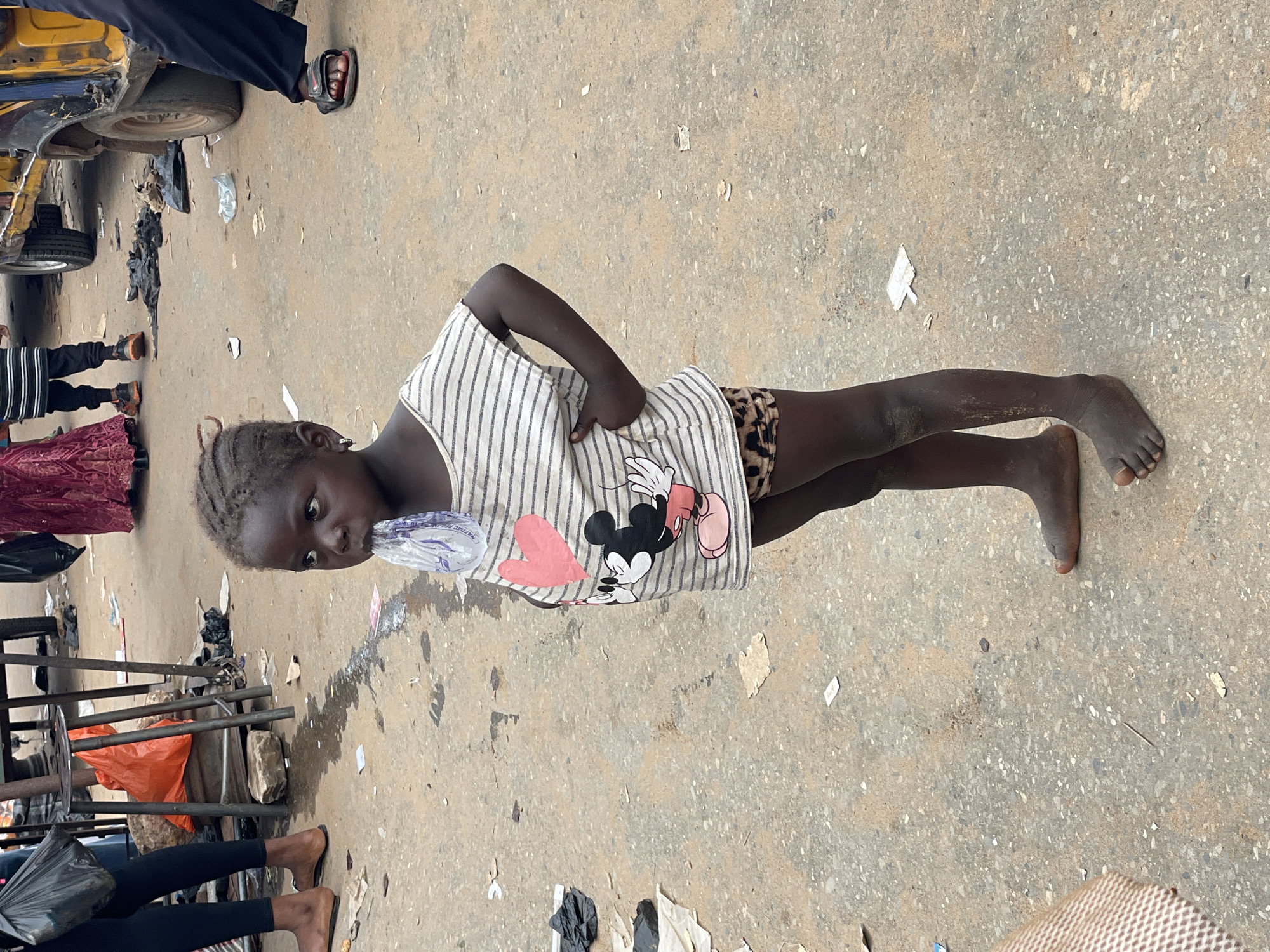 The image depicts an outdoor, paved area, likely a street or market in an unspecified city. The ground consists of grey asphalt or concrete, littered with scattered debris including crumpled papers, various plastic fragments, and several dark plastic bags. Dark liquid stains are present on the ground surface.
In the foreground, a young, dark-skinned child, estimated to be between 3 and 6 years old, stands barefoot. The child has dark hair styled in multiple small braids or twists. They are wearing a light-colored, horizontally striped t-shirt featuring a pink heart graphic on the left chest and a depiction of Mickey Mouse on the abdomen. Leopard-print patterned shorts cover the lower torso. The child holds a clear plastic sachet, containing a liquid, to their mouth, appearing to drink from it. Their right hand is placed on their hip, and their gaze is directed slightly upward and to the left.
In the mid-ground and background, multiple individuals are partially visible. To the left of the child, the lower legs and feet of an adult wearing dark pants and light brown sandals are seen, carrying a black plastic bag. Further left and behind, portions of metal-framed structures, possibly market stalls or tables, are present, with an orange plastic bag hanging from one.
To the right of the child, the lower section of a yellow and dark-colored motorized three-wheeled vehicle, resembling an auto-rickshaw, is visible, including its wheels and bodywork. Adjacent to the vehicle, the lower legs and feet of another adult, wearing dark pants and dark sandals with red accents, are present. In the far background, additional individuals are partially visible, including one person wearing a long, dark red patterned garment.