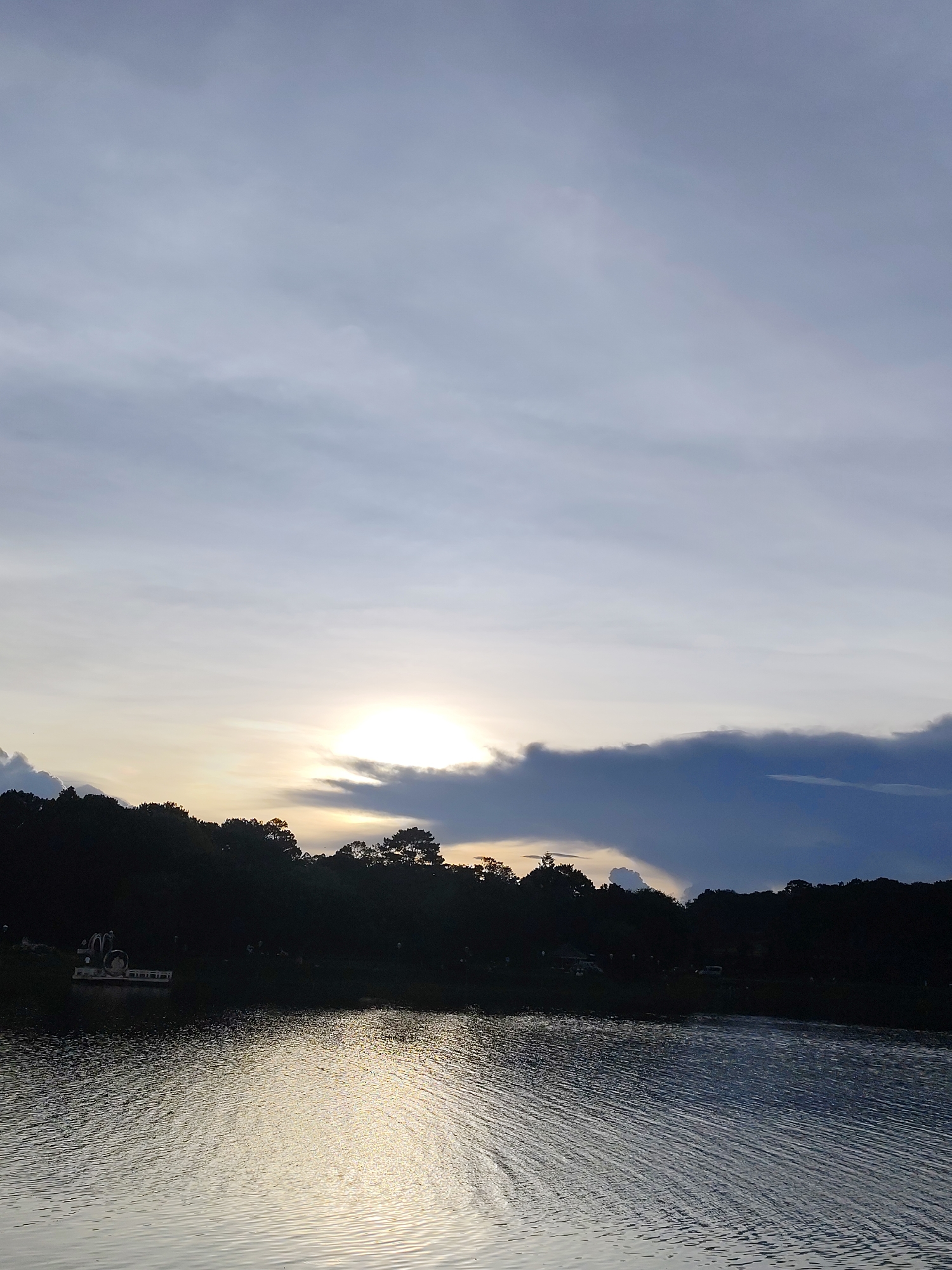A serene sunset over a lake in Da Lat, Vietnam, is captured in this vertical photograph. The sky, a canvas of pale blue and soft grey clouds, is dominated by the setting sun, a bright orb peeking through a gap in a dark, dramatic cloud formation. The sun's rays cast a shimmering golden path across the rippling surface of the lake, which occupies the lower half of the image. Silhouetted against the fading light, the far shore is lined with a dense forest of dark trees. On the left side of the shore, a white pontoon-like structure with a roof and what appears to be a decorative swan sculpture is visible. A few indistinct figures and structures are also faintly discernible along the treeline, suggesting human presence on the lakeside. The overall atmosphere is one of tranquility and natural beauty.