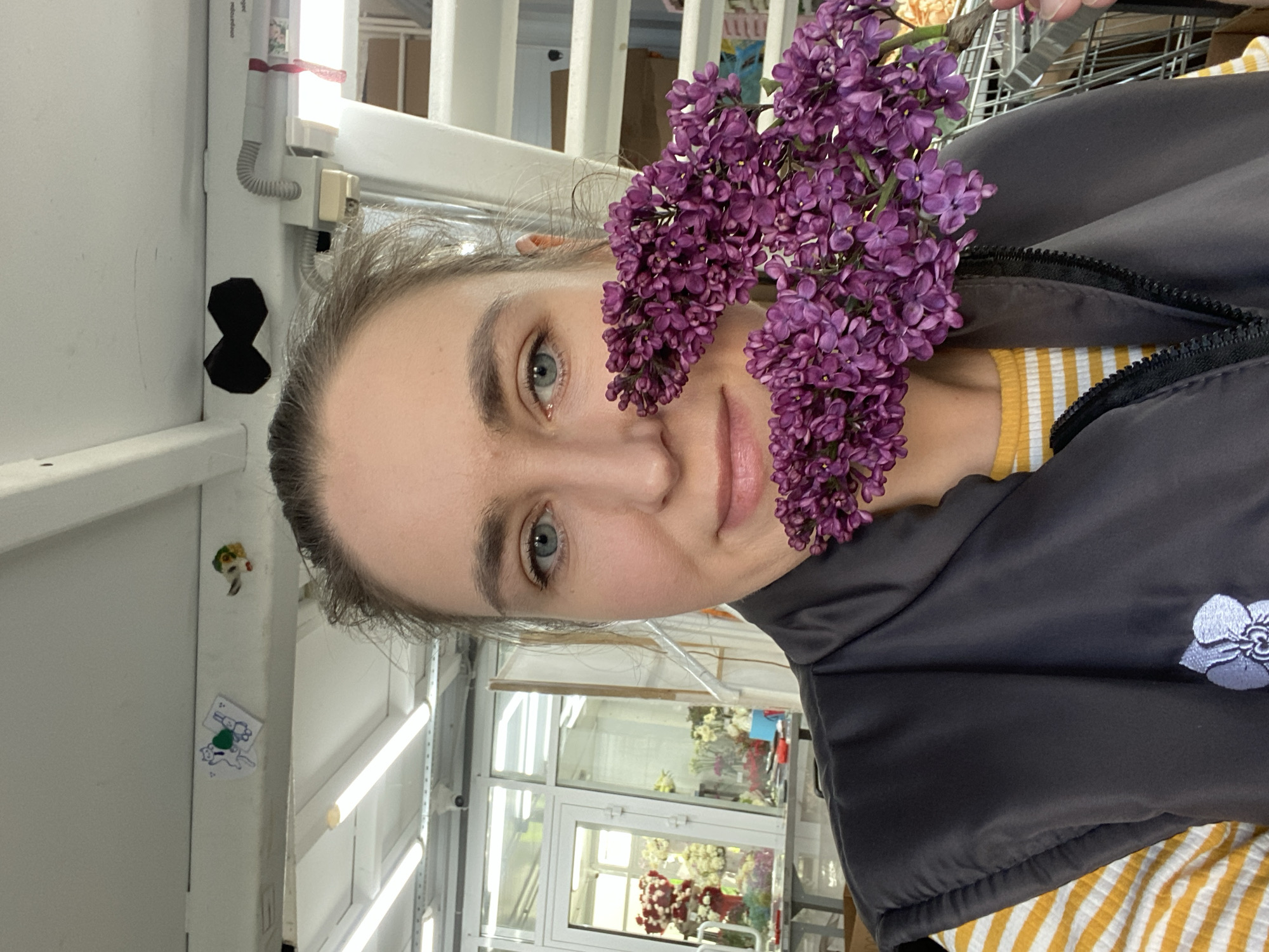A woman is holding a bouquet of purple flowers in front of her face. She is wearing a gray jacket over a yellow and white striped shirt. The scene appears to be set in a flower shop, which is located in Yekaterinburg, Russia. In the background, there are displays of flowers visible through a glass window.