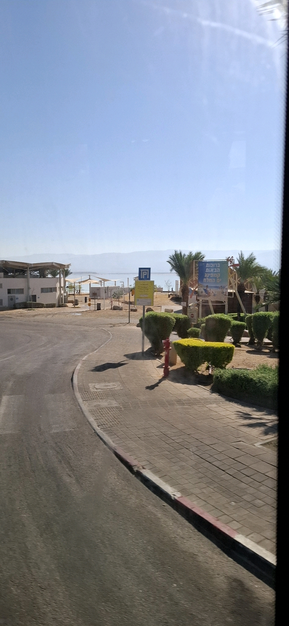 The image depicts a view of a paved area and a road leading towards a body of water, likely the Dead Sea, given the location in the Tamar Regional Council, Israel. On the left, a white building with a shaded canopy is visible. Further back, the Dead Sea stretches across the horizon, with mountainous terrain in the distance. The foreground features a paved walkway with red-and-white striped curbing and a paved road. Several manicured bushes and trees are strategically placed along the walkway. A red fire hydrant stands near a yellow and blue signpost. A large billboard with Hebrew text and images of people enjoying water activities is prominently displayed to the right. The sky is clear and blue, indicating a sunny day. No people are clearly identifiable in the immediate foreground or midground.