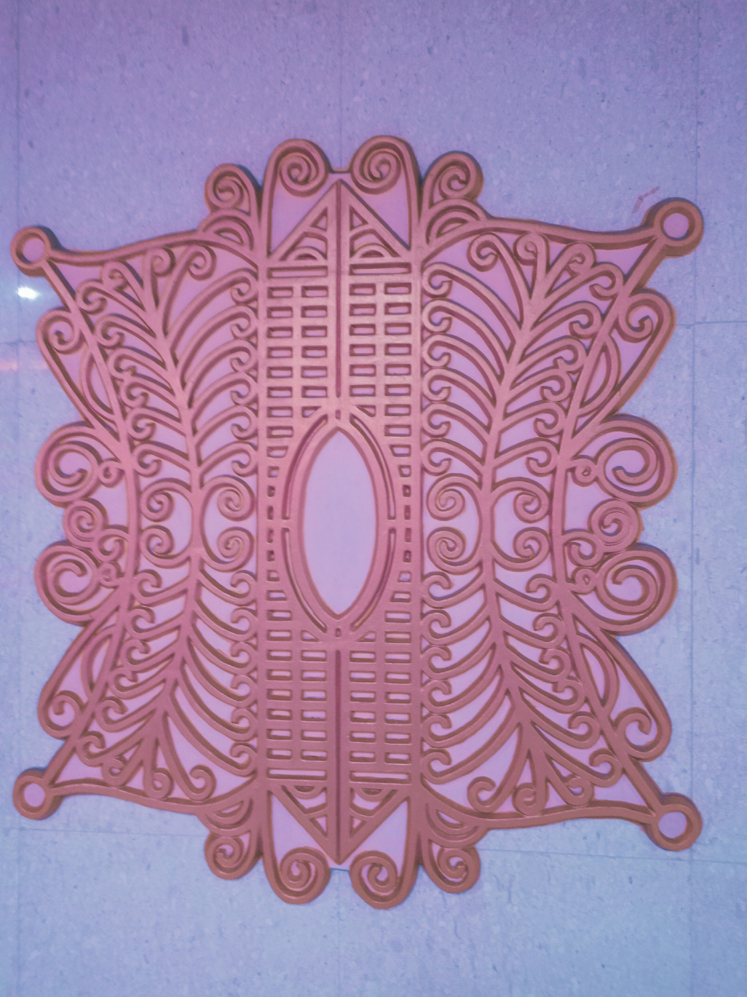 An eye-level close-up shot shows an ornate, symmetrical decorative element attached to a light-colored wall. The design is predominantly pink and gold, featuring a central oval shape flanked by intricate patterns resembling stylized leaves and scrollwork. The edges of the design are embellished with small, circular protrusions. The wall itself appears to be a textured, possibly tiled, surface. The city of Banda Aceh, Indonesia, is not discernible from the visual information in the image. There are no visible people or actions occurring.