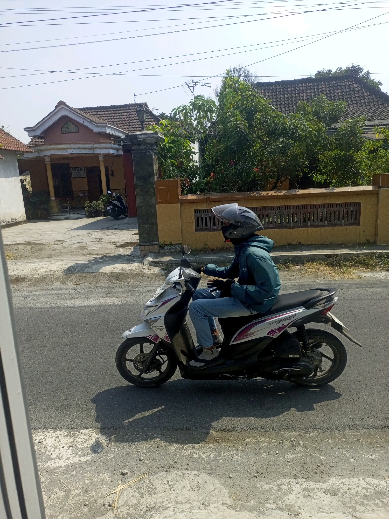 A person wearing a blue jacket and jeans rides a white and pink scooter down a paved road in Ngasem, Indonesia. The rider is wearing a grey helmet and black face mask. A house with a brown tile roof and a yellow wall with a decorative fence is visible in the background. The sky is clear and blue.