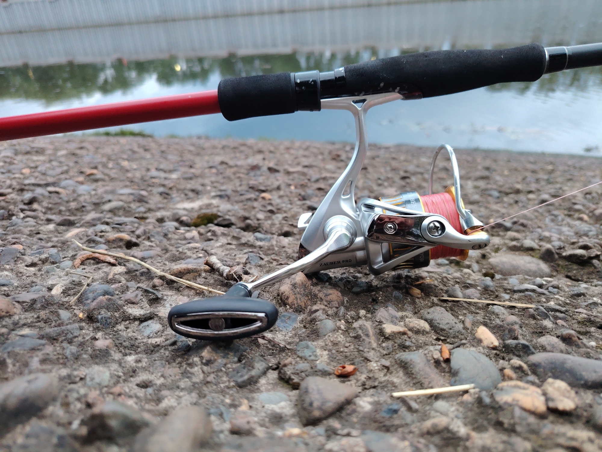 A fishing rod and reel lay on a stony surface, likely a riverbank. The fishing rod is red with a black foam handle. The reel is silver and black, with pink fishing line wound around the spool. The stony surface appears to be composed of small rocks and pebbles. A body of water is visible in the background, suggesting an outdoor setting. The location is not identifiable in this image.