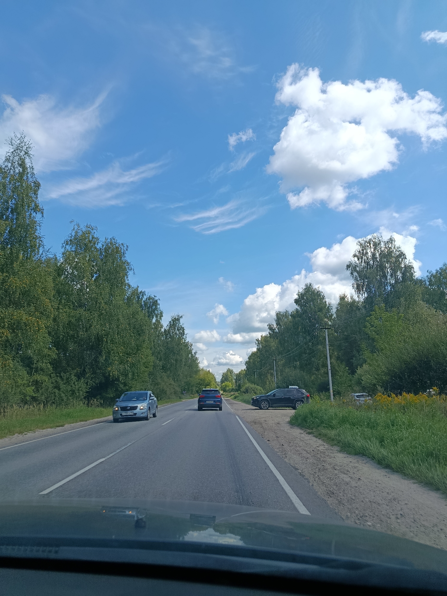 The image depicts a sunny day with a clear blue sky with scattered white clouds, viewed from inside a vehicle. The vehicle is on a two-lane asphalt road bordered by dense green trees and vegetation. There are at least three cars visible on the road: a light blue sedan in the left lane, a dark blue SUV in the right lane, and a dark SUV parked on the right shoulder of the road. A smaller silver car is also visible parked further down the right shoulder. Telephone poles are present along the right side of the road, interspersed within the trees. The environment suggests a rural or suburban setting, possibly on the outskirts of a city like Vereya, Russia, given the road infrastructure and surrounding natural landscape. The perspective indicates the image was taken from the driver's or passenger's seat of a moving vehicle.