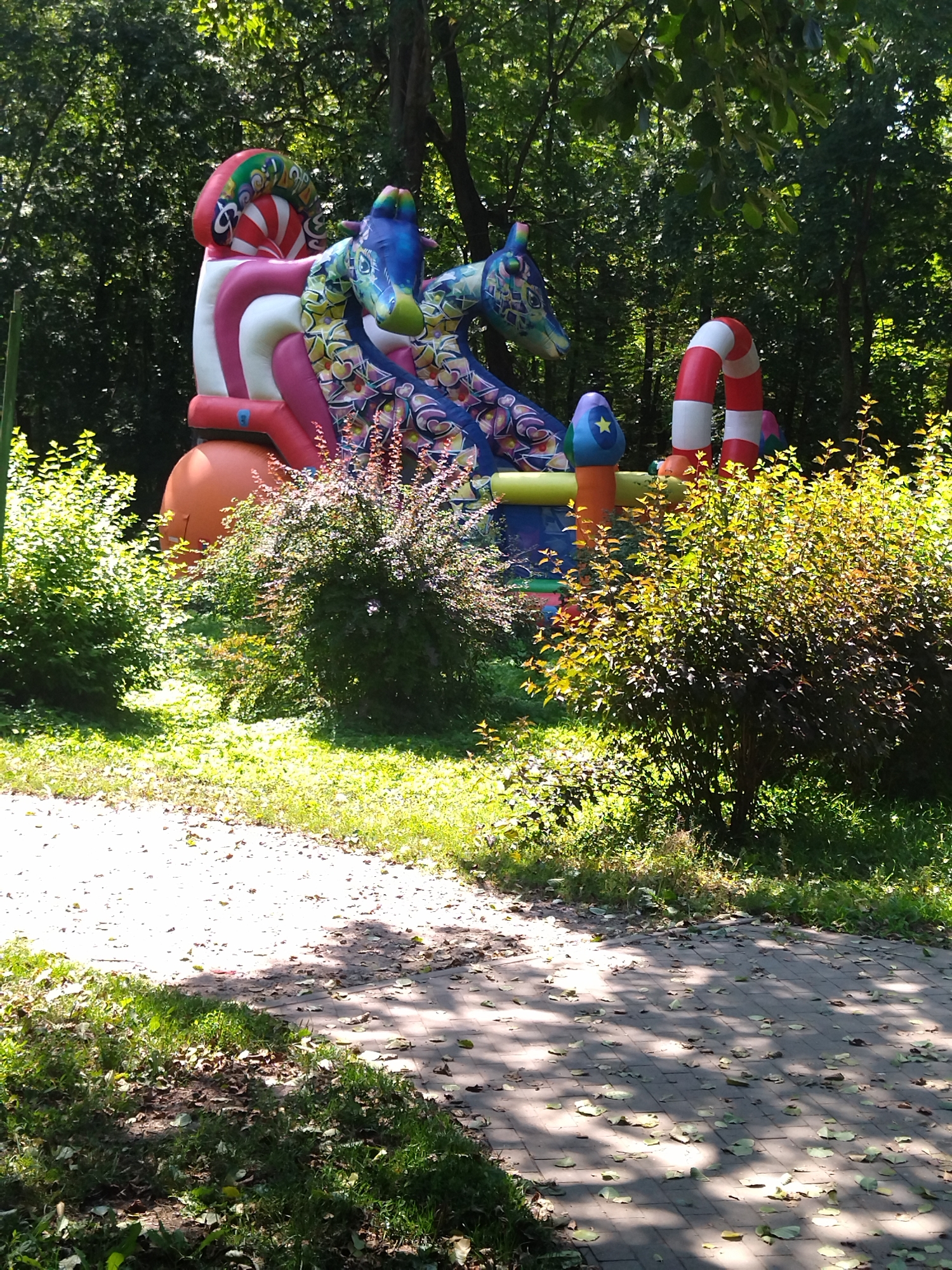 In Khmelnytskyi, Ukraine, an inflatable playground with a vibrant, multi-colored design, featuring two large, dragon-like creatures with intricate patterns, is situated in a park-like setting. The playground, primarily composed of red, white, pink, blue, and orange sections, includes a red and white striped arch. Surrounding the playground are lush green trees and bushes, casting dappled shadows on a brick pathway. The ground is covered with grass and fallen leaves, suggesting an outdoor recreational area. There are no people visible in the image.