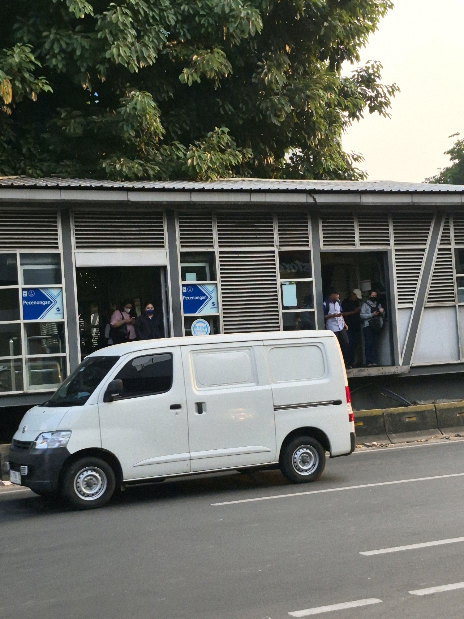A white commercial van is parked on the right side of the road in front of a modern bus stop in Central Jakarta, Indonesia. The bus stop features a long, covered structure with a sloped metal roof, supported by grey metal beams and framework. The walls of the bus stop are lined with horizontal louvers and large windows. Several people are visible inside and near the entrances of the bus stop, some wearing face masks. Blue signage with white text, including the word "Pecenongan," is displayed on the exterior of the bus stop. The road in the foreground is asphalt with white lane markings. Trees with dense green foliage are visible above and behind the bus stop structure. The sky is overcast with a pale yellow hue, suggesting either early morning or late afternoon.