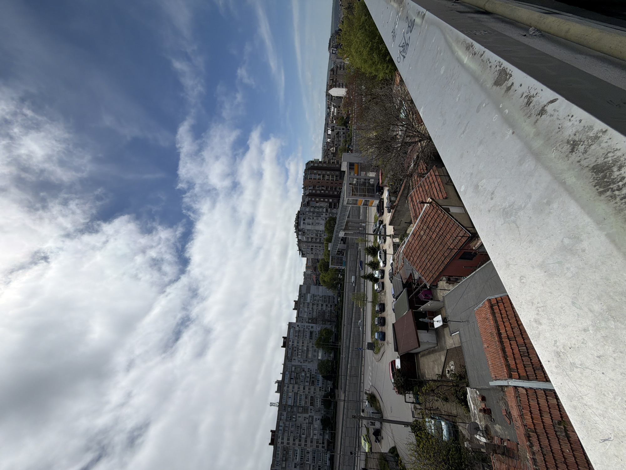 An elevated, rotated perspective shows a cityscape in Varna, Bulgaria. The sky occupies the left of the frame, showing patches of blue and white clouds. The city below includes apartment buildings, houses with red-tiled roofs, a road with parked cars, and some trees. A gray concrete structure is located on the top right of the image.