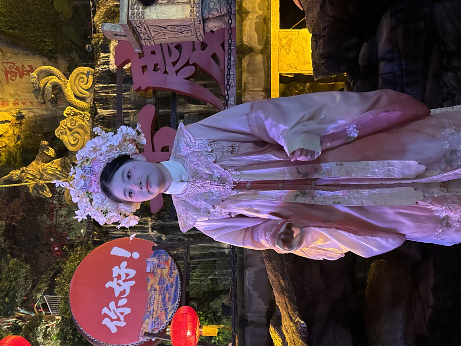 A young female stands centered in the frame, dressed in elaborate traditional Chinese attire, illuminated at night in Yuzhong District, China. She wears a multi-layered pink and white robe featuring embroidered floral patterns, a prominent decorative collar with hanging embellishments, and a white collared inner garment. Her head is adorned with a large, elaborate headpiece composed of numerous white and light pink artificial flowers. Subtle pink markings are visible on her cheeks. Her posture is upright, with her hands partially visible, held in front of her body. She looks slightly upward and to her left with a neutral expression.
The background consists of a multi-tiered rock-like structure with water flowing over it, resembling a waterfall. On the upper left, a large red circular sign displays white Chinese characters "你好!" (Hello!), with a red traditional lantern hanging below it. Above the central waterfall, golden sculptures are visible, including a standing figure holding a staff and a stylized, two-headed serpent or dragon. Large red Chinese characters are partially visible on the right side of the background structure. The foreground contains dark, uneven rock formations, some illuminated by yellow light from below. The overall environment suggests an elaborately themed outdoor setting.