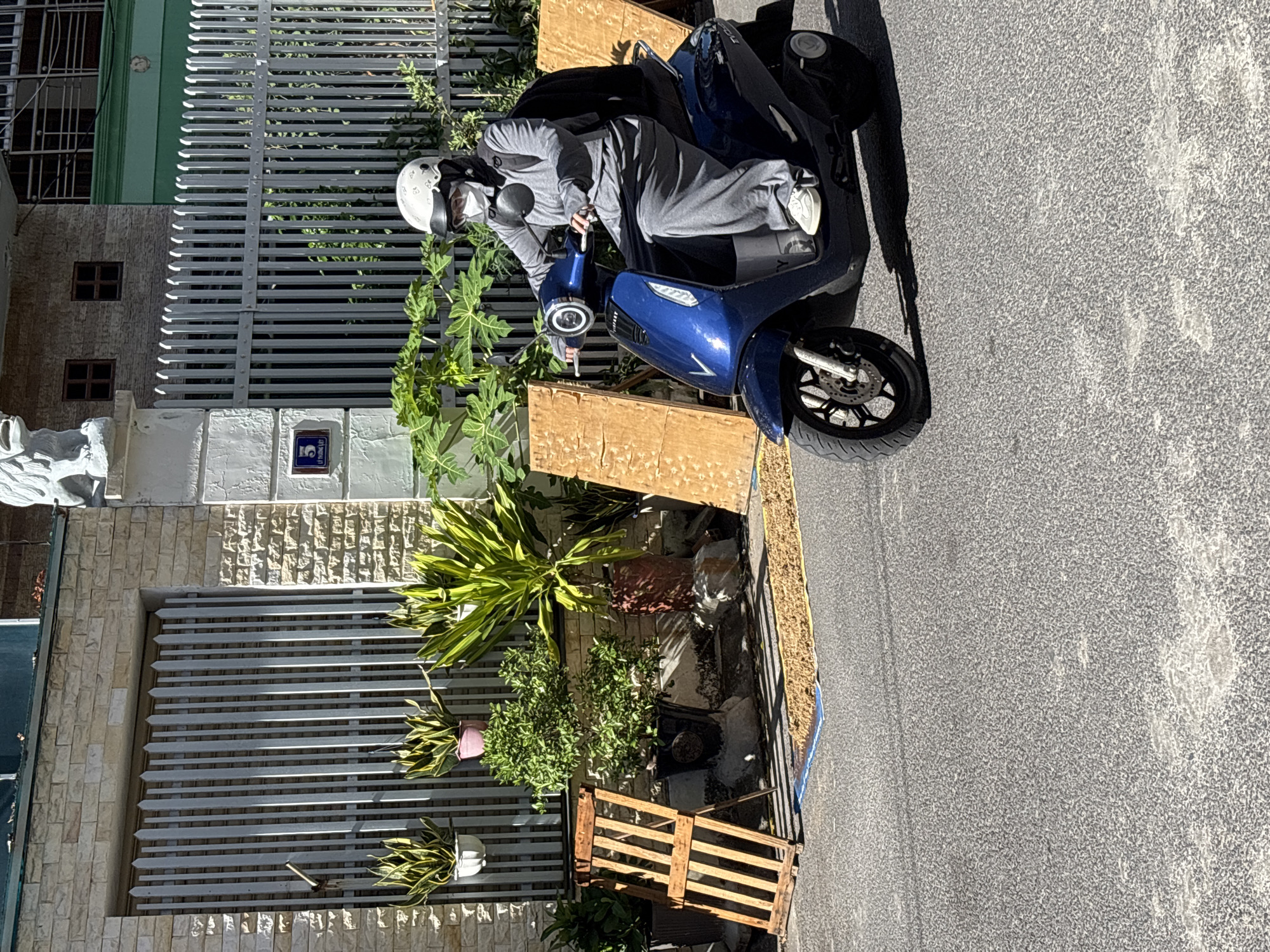 A person wearing a white helmet, face mask, and a gray hooded outfit rides a dark blue electric scooter on an asphalt road. The scooter has a black backpack on the back. The person is wearing gray pants and white sneakers. The scooter is a VinFast brand, as indicated by the logo on the side.
In the background, there is a building with a stone facade and a tall metal gate with vertical slats. Several potted plants are placed in front of the gate, including some hanging plants. To the right of the scooter, two large, rectangular wooden panels are leaning against a structure. A strip of blue tarp with a scattered brown material on top is visible on the ground in front of the wooden panels.
The scene appears to be a street in Nha Trang, Vietnam, given the presence of a numbered sign with Vietnamese text ("LY THUONG KIET 5") on the gate post. The building has architectural elements suggestive of a residential or commercial property in the region, and the scooter is a common mode of transportation in Vietnamese cities. The overall impression is of a typical urban street scene on a sunny day.