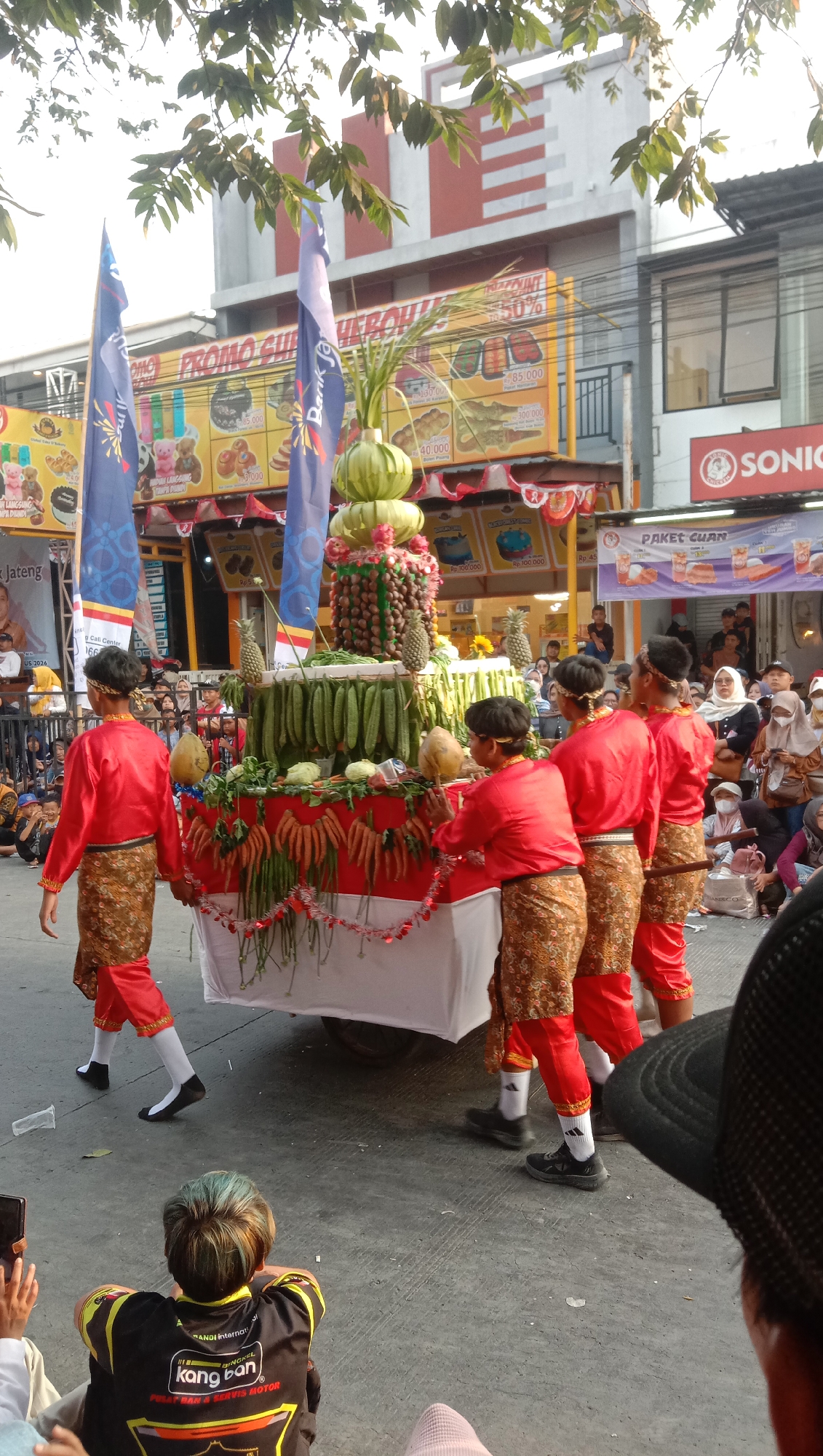 A parade is underway in Jatisawit, Indonesia, featuring a large, mobile altar adorned with various fruits and vegetables. The altar is decorated with red and white fabric, resembling the Indonesian flag, and is surrounded by a garland of white and red tassels. Several young men, dressed in traditional red shirts and patterned trousers, are actively participating in the parade, appearing to push or guide the altar. The procession is taking place on a paved street lined with buildings and banners advertising local businesses and promotions. A crowd of onlookers, some wearing face masks, watches the parade from the sidewalks. The scene is illuminated by natural sunlight, casting shadows and highlighting the vibrant colors of the decorations and attire. The overall atmosphere suggests a festive cultural event.