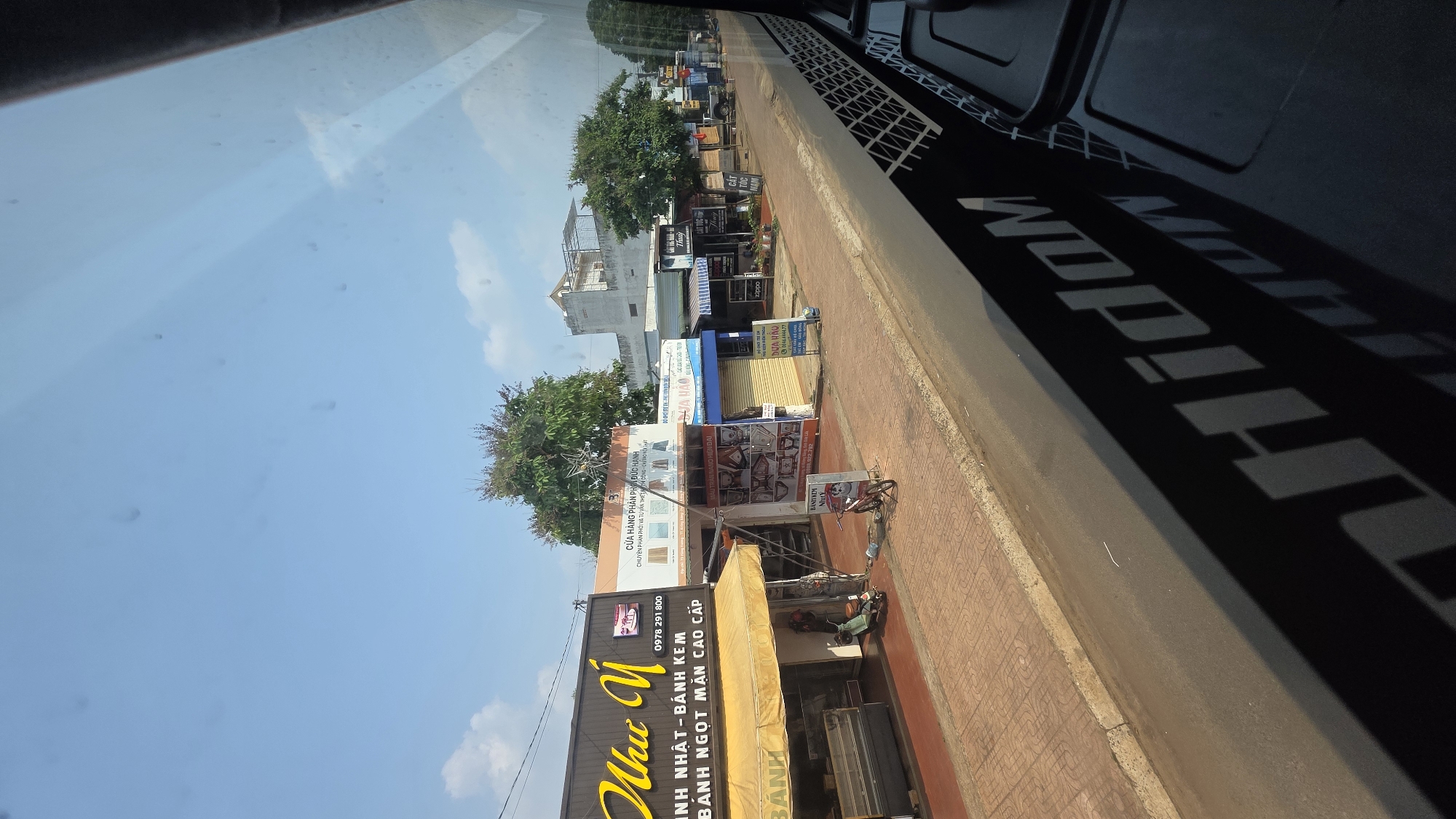 Captured from inside a vehicle, the image portrays a street scene in Xuân Long, Vietnam. The camera is positioned high, offering a view out the window and a portion of the vehicle's interior. Outside, a clear blue sky speckled with wispy clouds meets the rooftops of closely packed buildings lining the street. The businesses display signs in Vietnamese, advertising a variety of goods and services, including baked goods, home furnishings, and hair salons. Bicycles and scooters are parked on the sidewalk in front of the shops. The vehicle's interior, visible in the foreground, includes part of the dashboard. The lighting is bright, suggesting a sunny day.