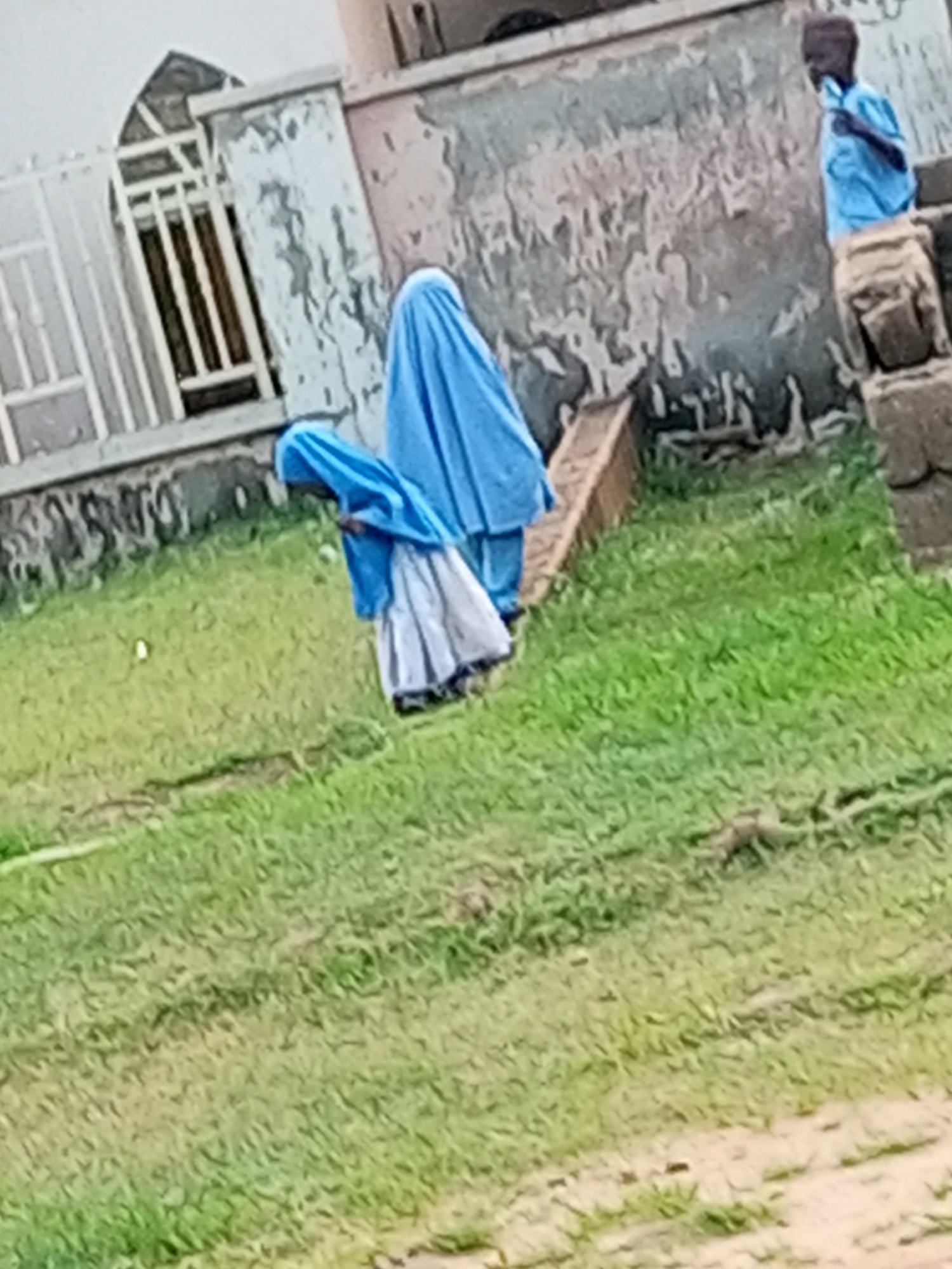 In Tempure, Nigeria, three individuals are visible in front of a dilapidated building. On the right, a young boy wearing a blue shirt stands facing left, looking downwards. In the center, a person wearing a bright blue hijab and a white skirt bends over. To their left, another person also wears a bright blue hijab, with a glimpse of a white skirt visible. They appear to be interacting. The background features a weathered, light-colored wall with visible cracks and peeling paint. To the left of the building, a white metal fence with a peaked design is present. The foreground consists of a grassy area with patches of bare earth and some debris.