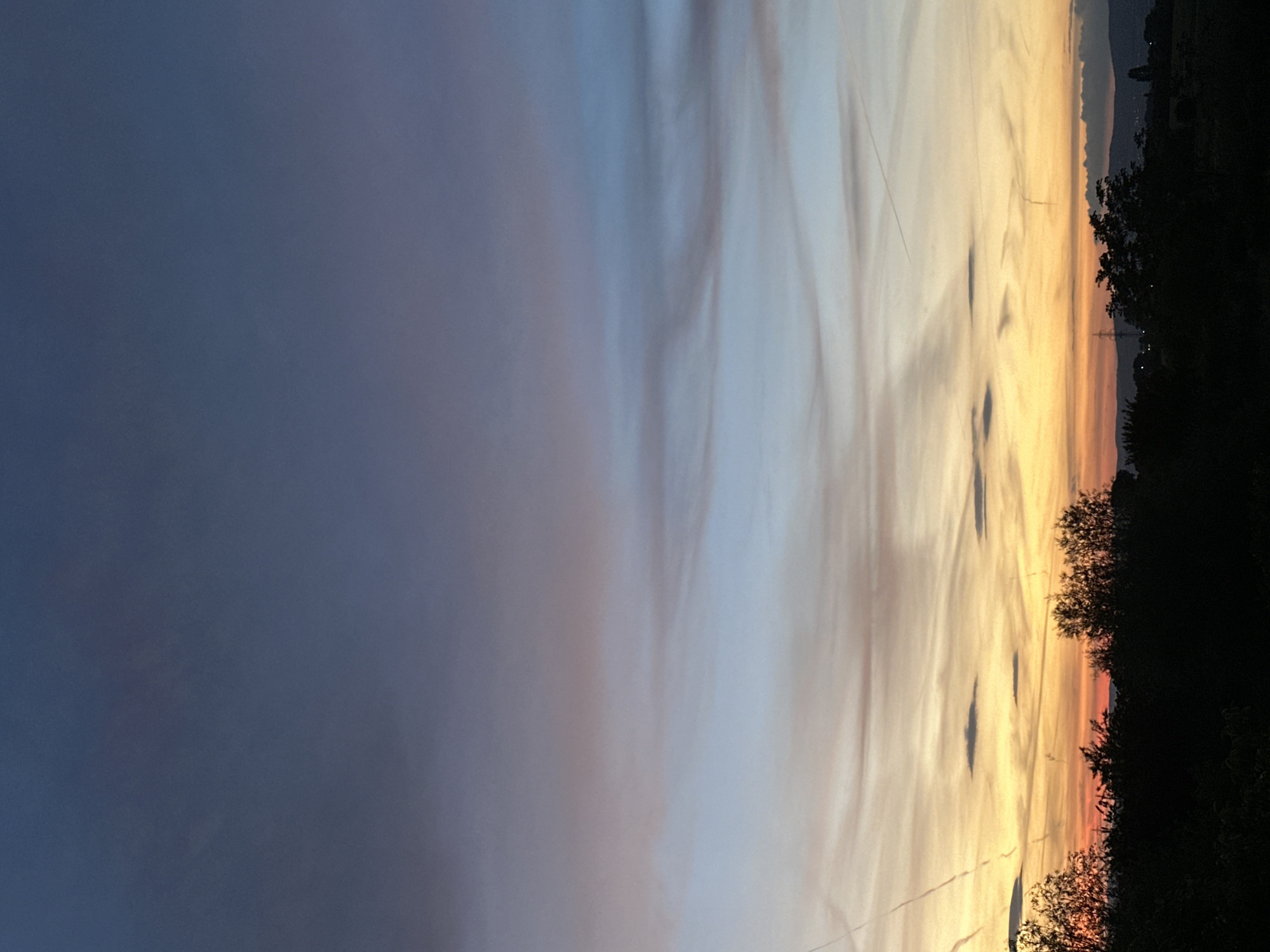 The image captures a sunset view. The sky transitions from a deep blue on the left to orange and yellow hues on the right horizon. Wispy clouds are scattered across the sky. A dark treeline is visible on the right, with silhouettes of trees against the vibrant sky. There is no specific information about location in Sofia, Bulgaria.