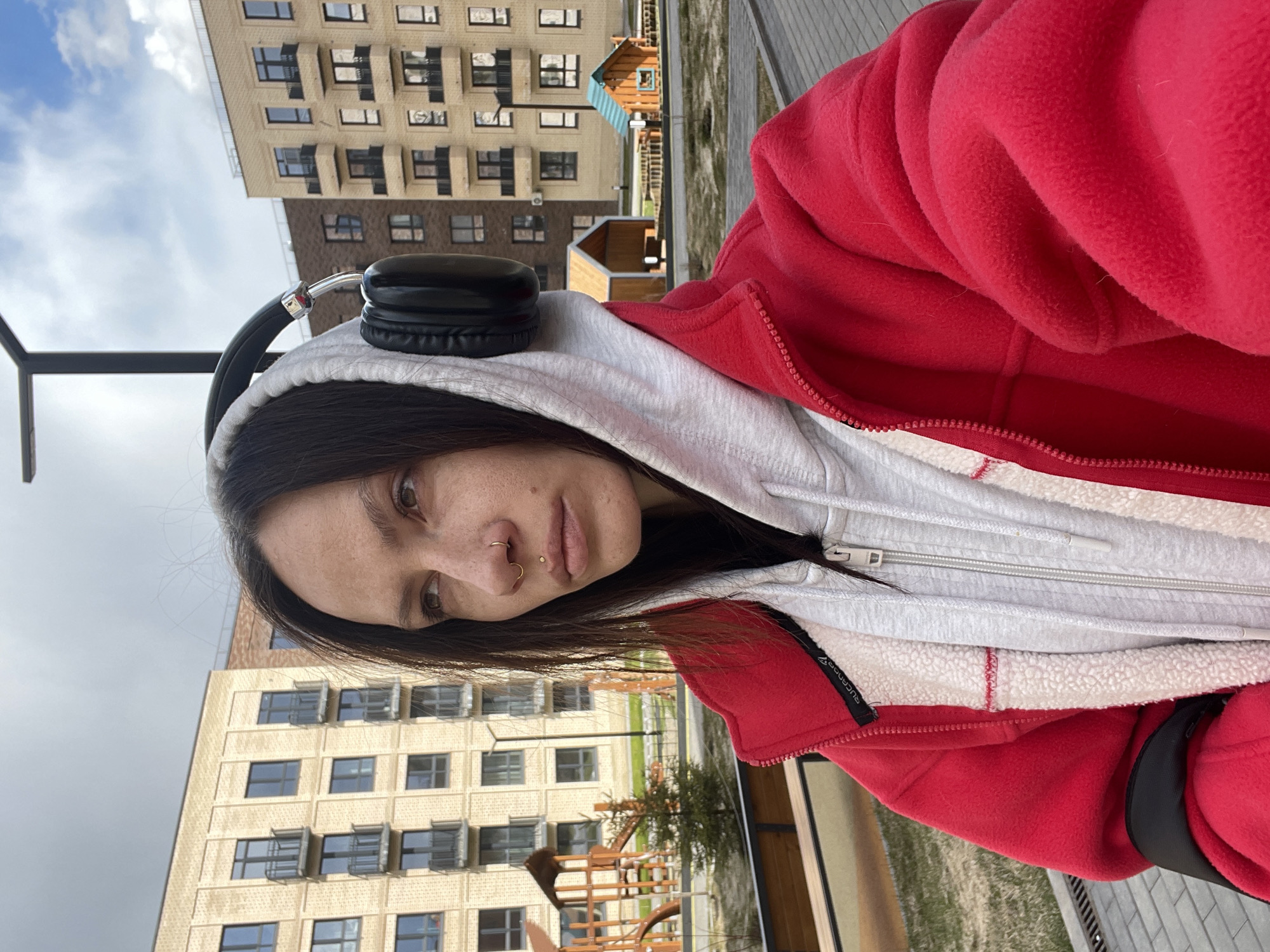 A selfie captures a young woman outdoors in a residential area of Izhevsk, Russia. She looks directly at the camera with a neutral or slightly pensive expression. Her dark hair frames her face, revealing freckles and brown eyes. She wears several facial piercings: a gold hoop on her septum, a small gold stud on her left nostril, and a gold stud on her philtrum. Black over-ear headphones are perched on her head. Her attire consists of a light grey zippered hoodie layered under a bright red fleece zippered jacket. A black tag with "SUCANOOZ" in white text is visible on the red jacket's inner lining.
The background shows multiple modern, multi-story apartment buildings, appearing to be constructed from light-colored brick or siding, with numerous windows, some featuring external shade structures. Between the buildings, a children's playground with wooden structures is visible, alongside patches of green grass and trees. The sky above is largely overcast with some lighter clouds, indicating it is daytime, possibly on a cool day given the woman's warm clothing. The scene suggests a communal space within an urban or suburban housing complex.