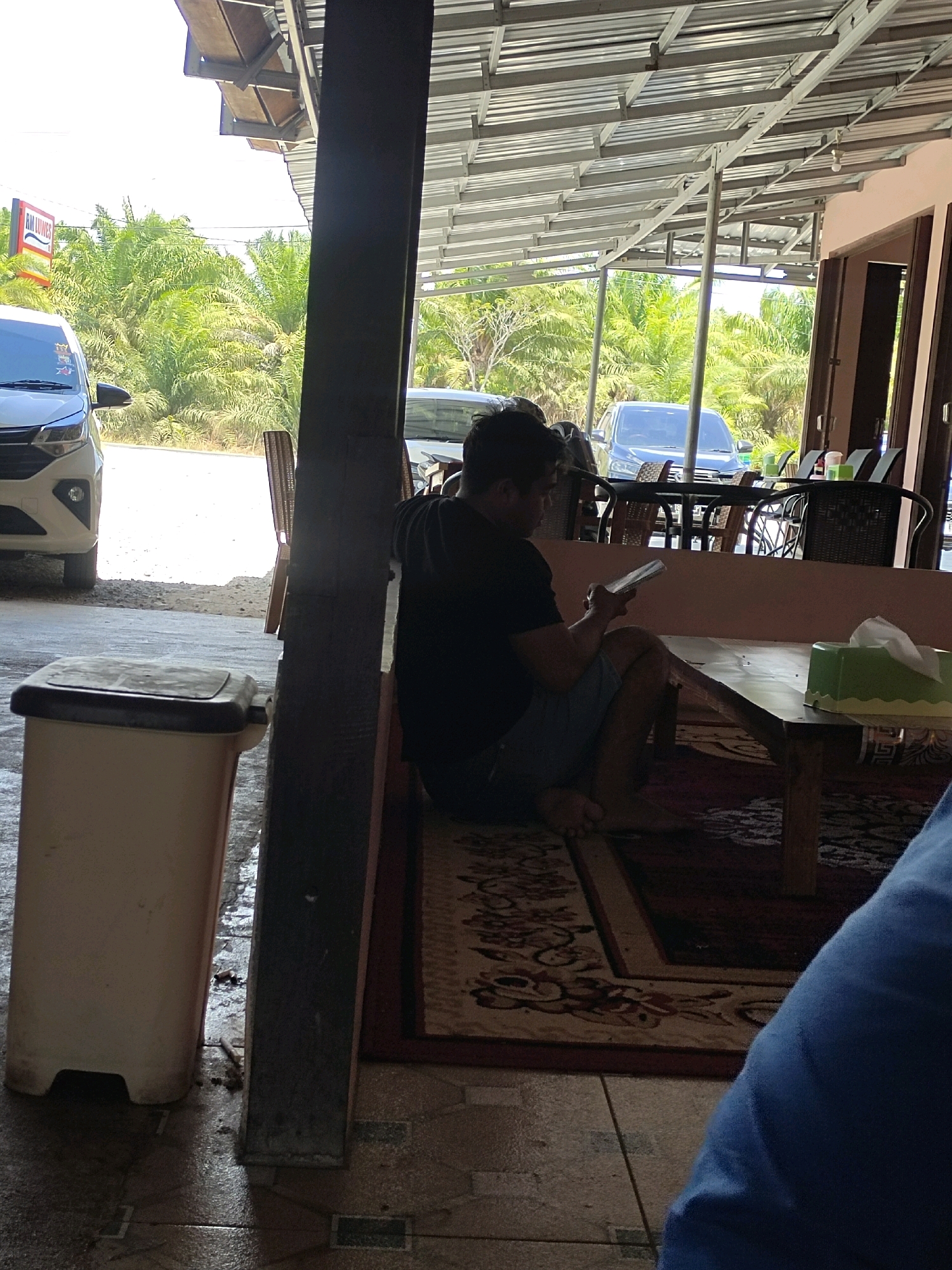 In Jilatan, Indonesia, a man sits on the floor with his legs crossed, engrossed in a mobile device. He is seated on a rug in what appears to be an outdoor or semi-outdoor seating area, likely a restaurant or roadside stop. The area is covered by a metal roof supported by pillars, and palm trees are visible in the background, suggesting a tropical or subtropical environment. A white trash can is positioned to the left of the man. In the foreground, a portion of a blue fabric is visible, possibly clothing or a blanket. The overall scene conveys a relaxed, informal atmosphere.