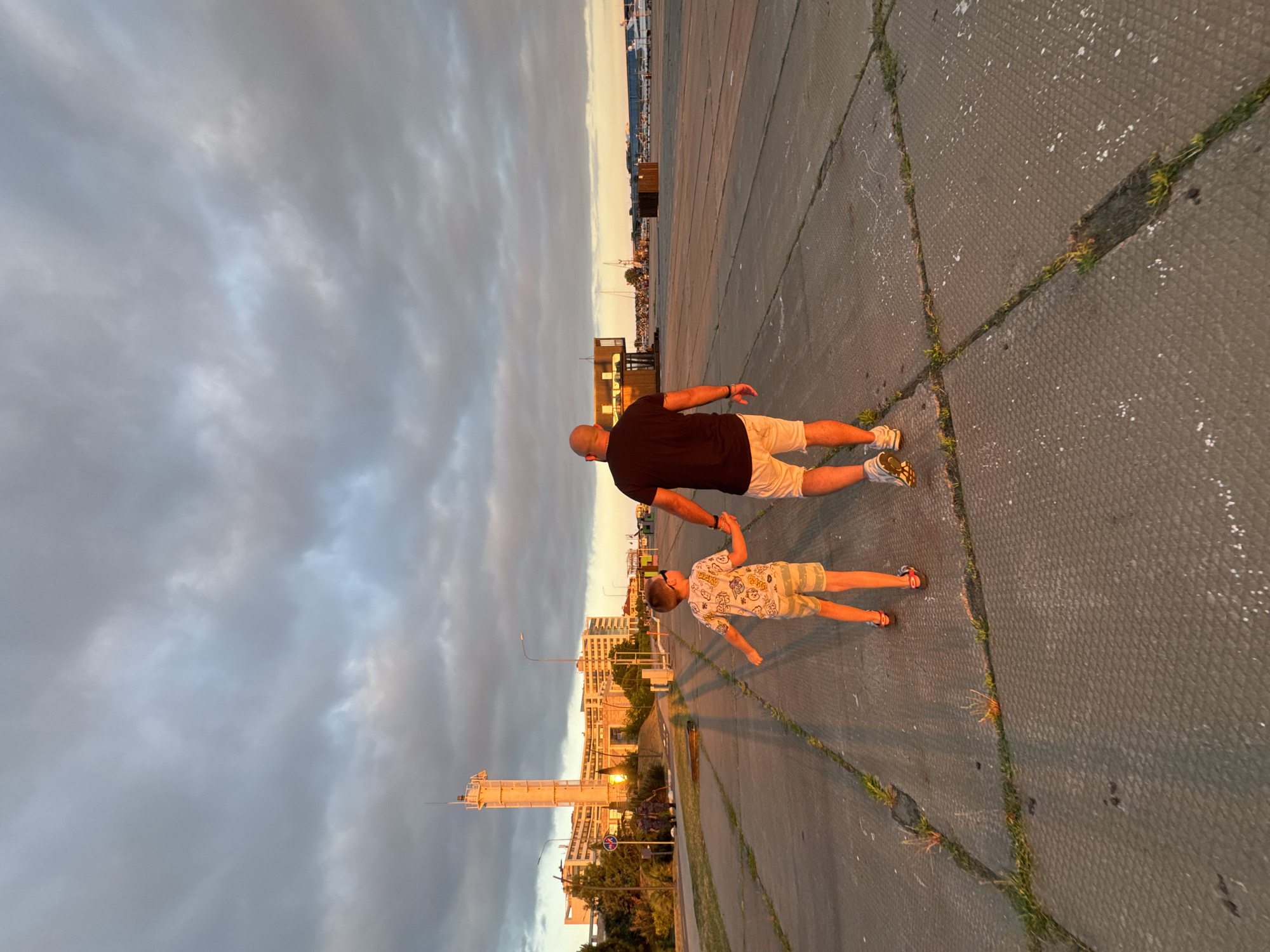 The image captures a father and son walking hand-in-hand on a paved walkway, likely in the city of Sirius, Russia, during sunset. The low angle of the sun casts long shadows across the textured concrete ground, illuminating the scene with a warm, golden light. In the background, a tall, slender tower, possibly a radio tower or lighthouse, stands prominently against a sky filled with dramatic, gray clouds that are tinged with the hues of the setting sun. Adjacent to the tower is a modern, multi-story building with numerous windows, reflecting the light. Further in the distance, a harbor or port area is visible, with numerous boats and a long, low building. The overall atmosphere is one of peacefulness and familial connection against an urban, coastal backdrop.