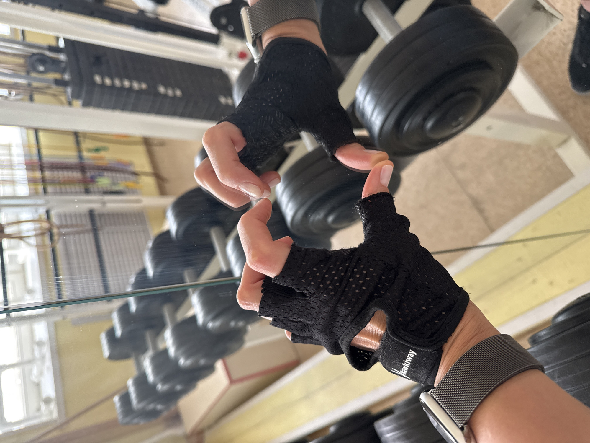 The image shows a person's hands, wearing fingerless black workout gloves and a gray metal mesh watchband, touching their reflection in a mirror. The reflection shows the hands and glove clearly, along with a blurred background. The background includes a weight rack with black weight plates. The location is not definitively identifiable as Chaikovsky, Russia, based on visual information alone.