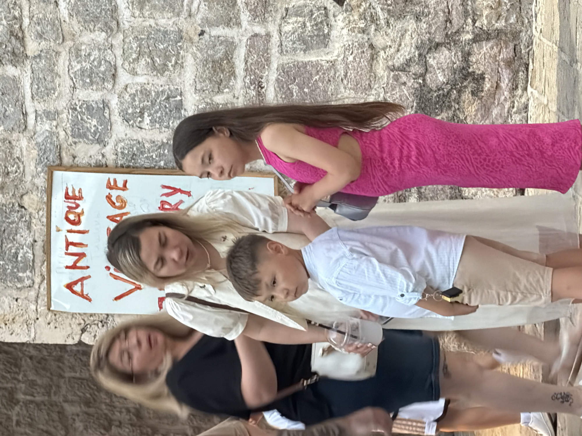 The image shows a group of people standing in front of a stone wall with a sign that reads "ANTIQUE VINTAGE GALLERY". A woman in a black top and a white purse is on the left. To her right is a woman in a cream-colored dress holding a drink. A young boy in a white shirt and beige shorts is in the foreground. To the right of the boy is a young girl in a bright pink dress. The background is a textured stone wall. The setting appears to be an outdoor market or street in Budva, Montenegro, given the architecture and the likely context of an "Antique Vintage Gallery" sign.